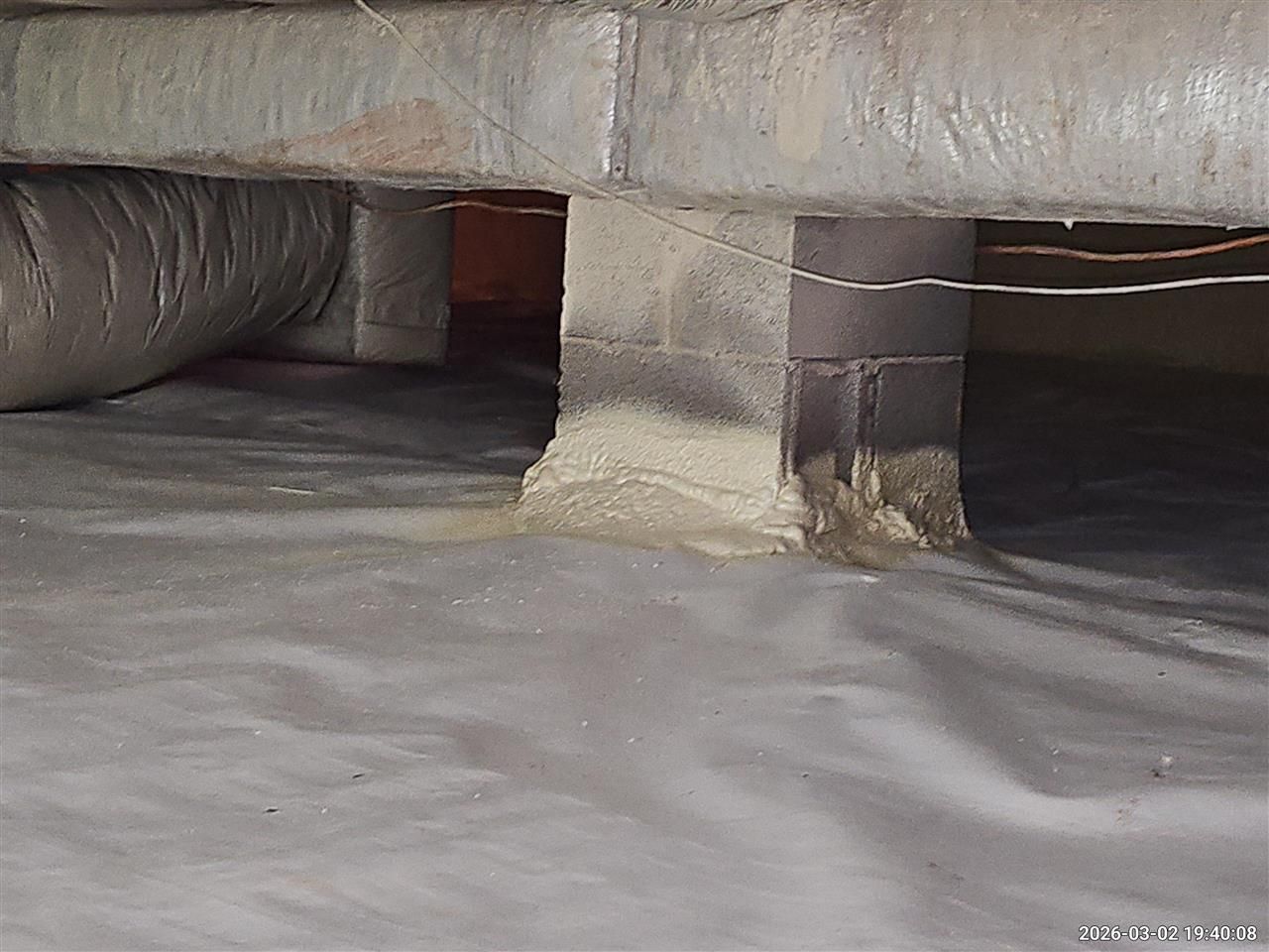 A concrete pier support for a house foundation stands on a white vapor barrier in a crawl space.