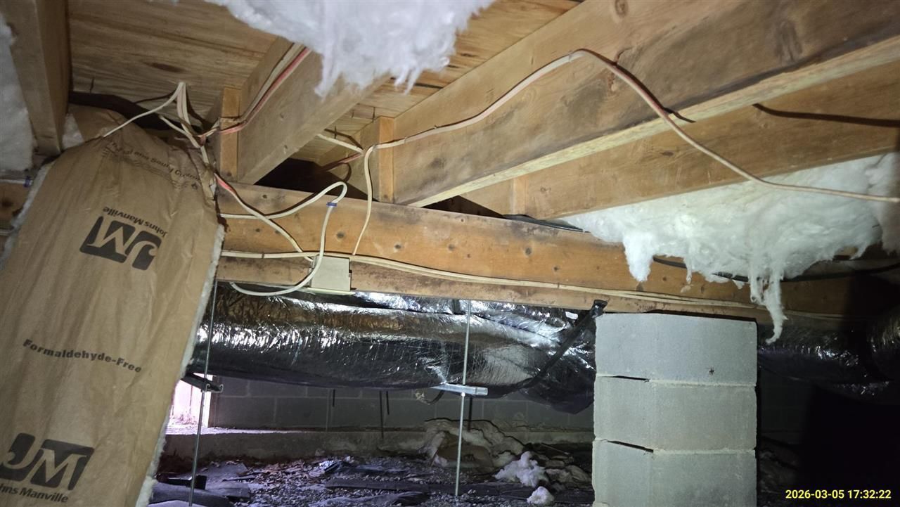 Crawlspace with exposed wooden floor joists, hanging fiberglass insulation, white foam sealant, and wiring.
