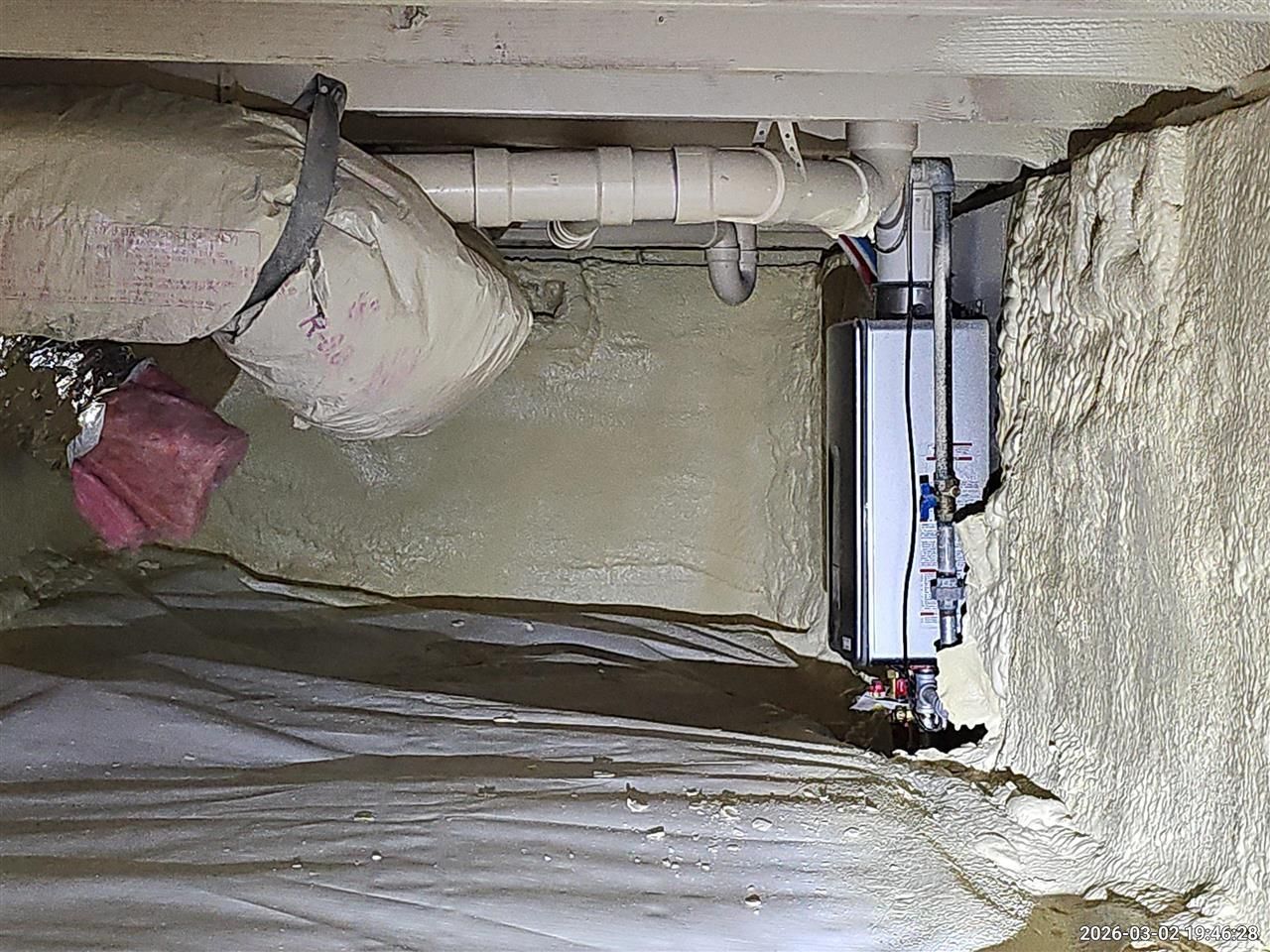 A tankless water heater mounted on a spray-foamed foundation wall in a crawl space with a vapor barrier floor.