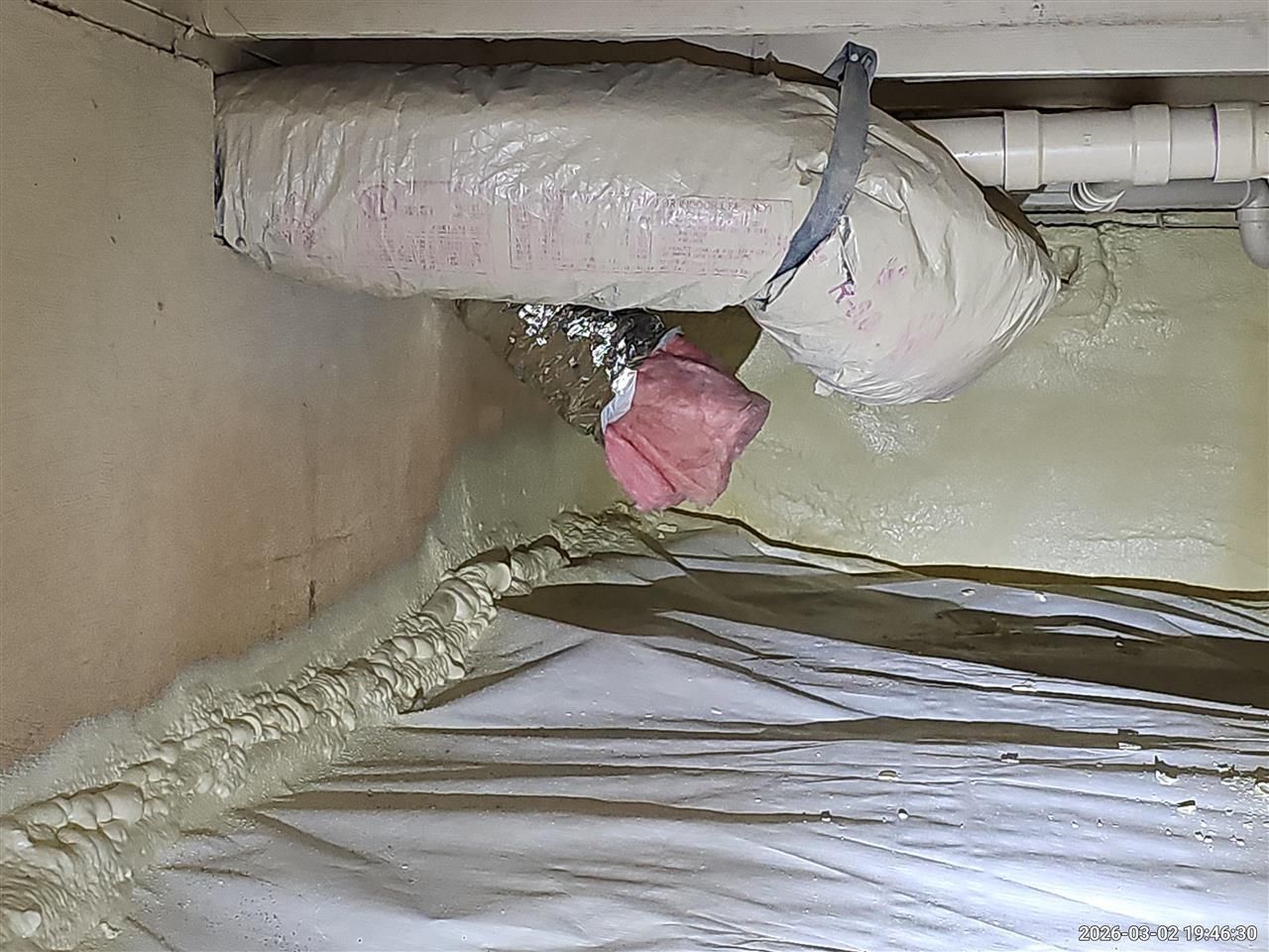 An insulated duct suspended in a crawlspace with white vapor barrier plastic on the ground and foam-sealed wall seams.