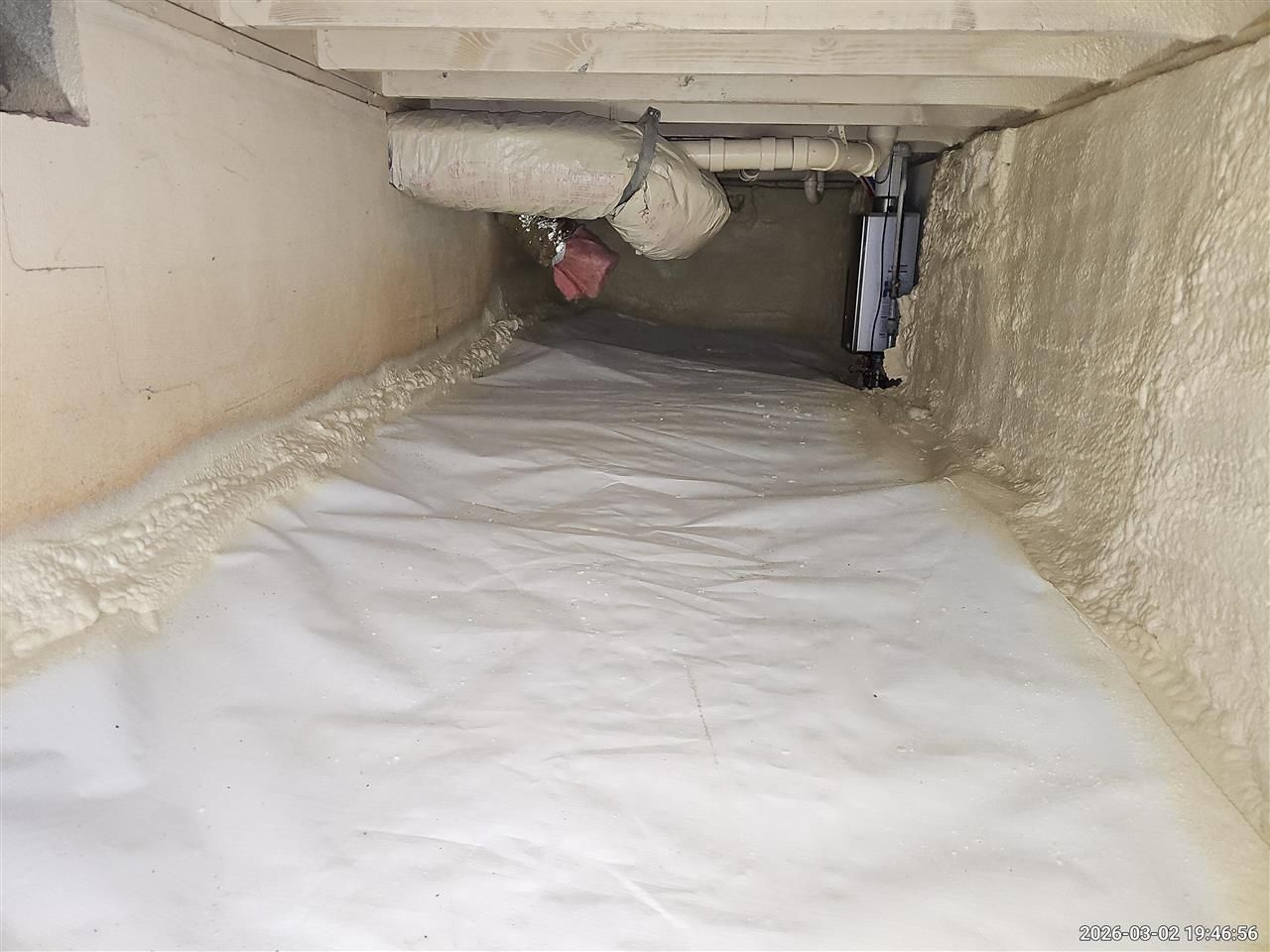 A white vapor barrier lines the ground of a crawl space with spray foam insulation on the foundation walls.
