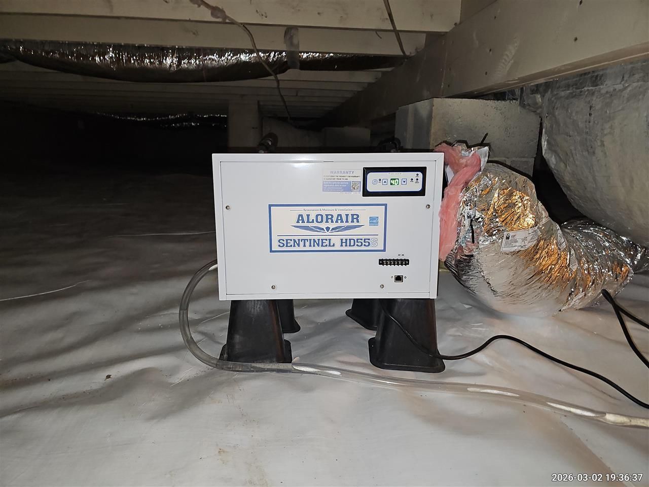 A white Sentinel dehumidifier unit sits on black support stands on a white vapor barrier in a crawl space.