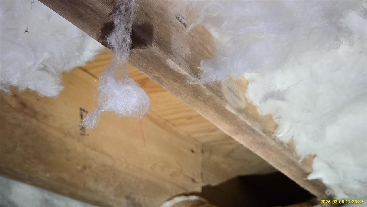 A close-up view of fiberglass insulation hanging from a wooden beam near a dark gap in a building structure.