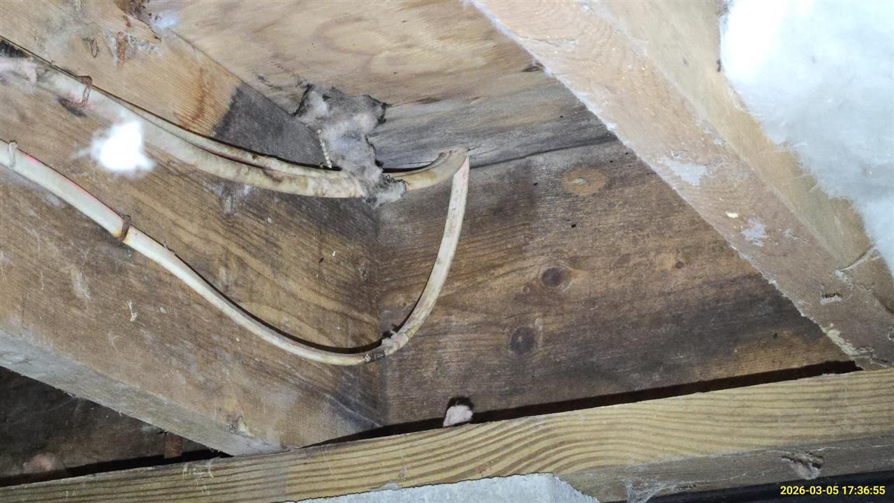 An attic space with exposed wooden rafters, white electrical wiring running along a beam, and patches of gray insulation.