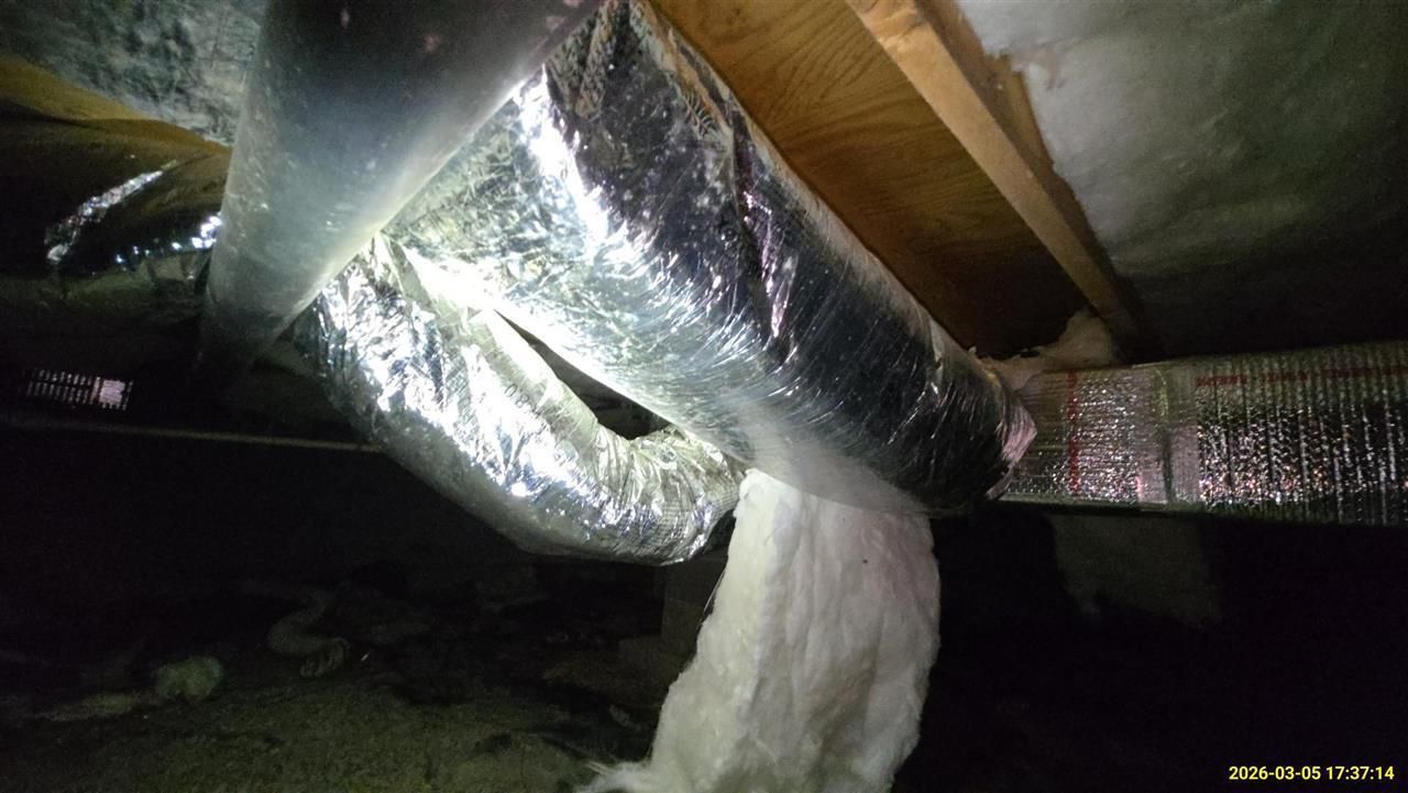 Insulated HVAC flexible ductwork hanging from floor joists in a dark crawlspace with exposed fiberglass insulation.