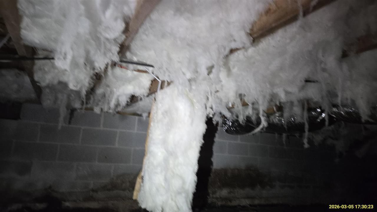 White fiberglass insulation hangs in disarray from the floor joists of a dim, concrete-block crawl space.