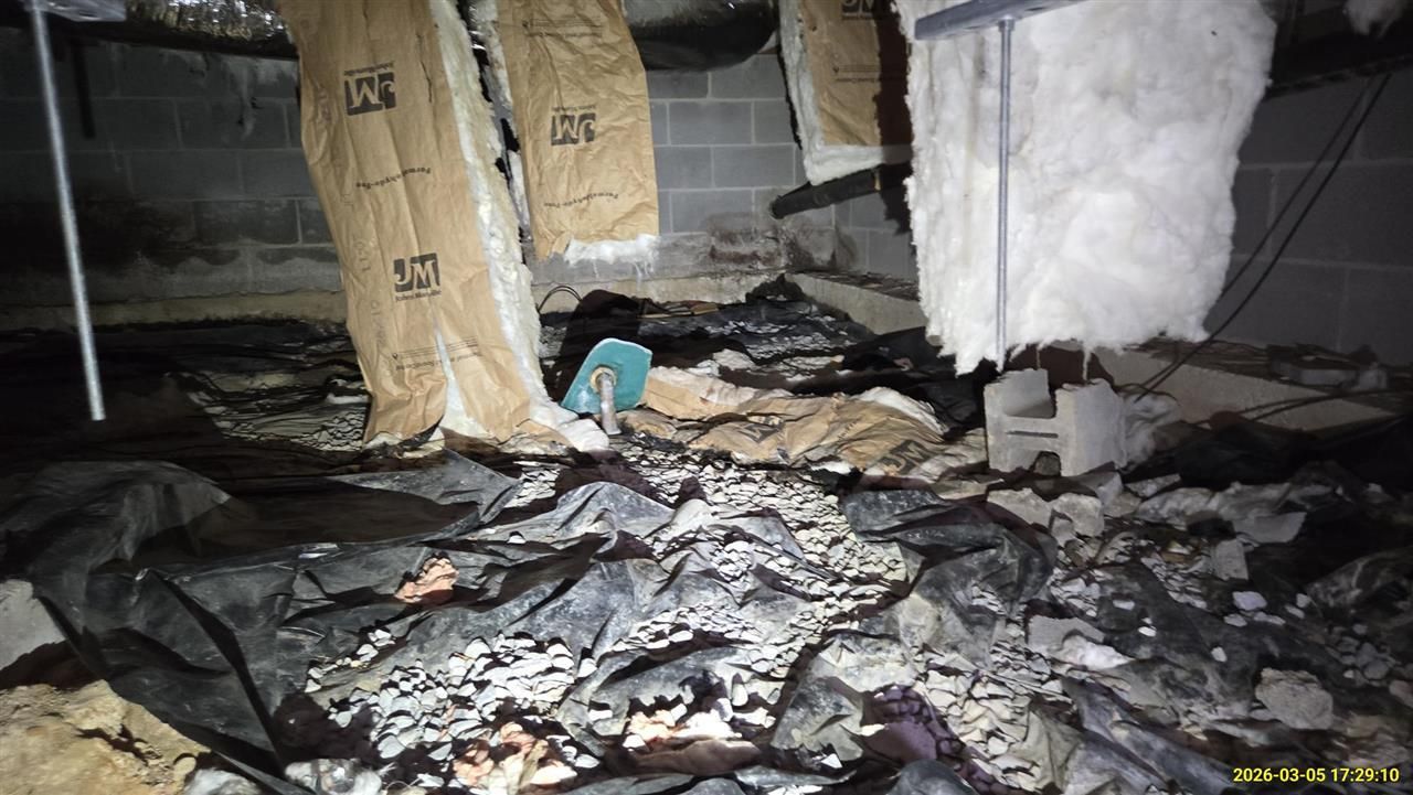 A dark crawl space with sagging insulation hanging from floor joists above a torn, debris-covered black vapor barrier.