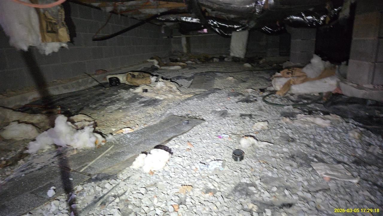 A dimly lit crawl space showing debris, fallen insulation on the ground, and exposed ductwork attached to floor joists.