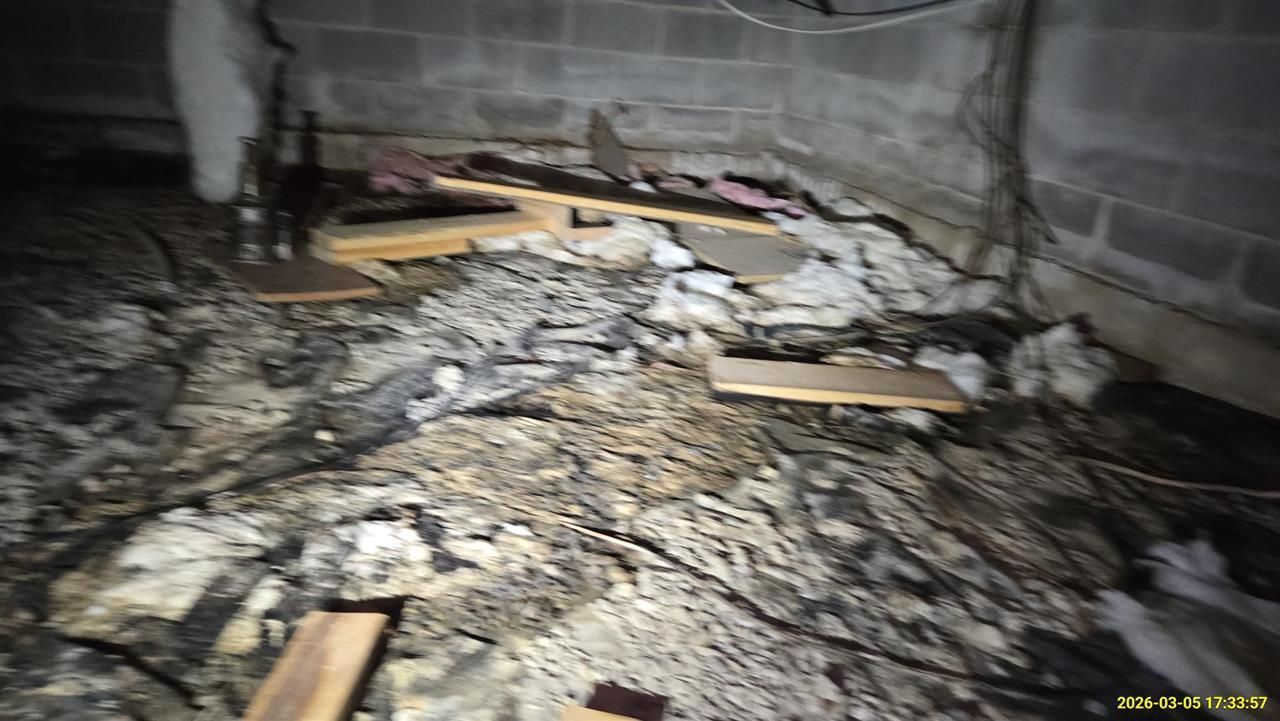 A dark, damp crawlspace corner featuring concrete walls, scattered wooden debris, and patches of wet insulation.