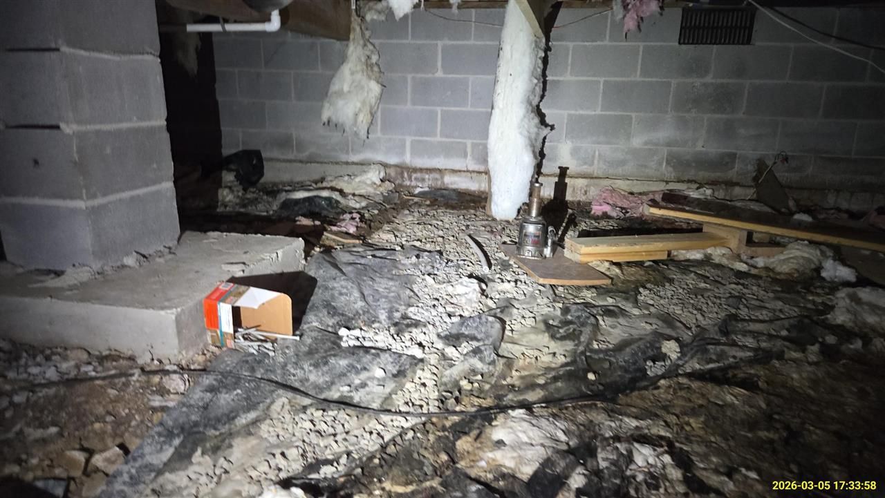 A dark, cluttered crawlspace with concrete blocks, hanging insulation, and debris scattered across the ground.