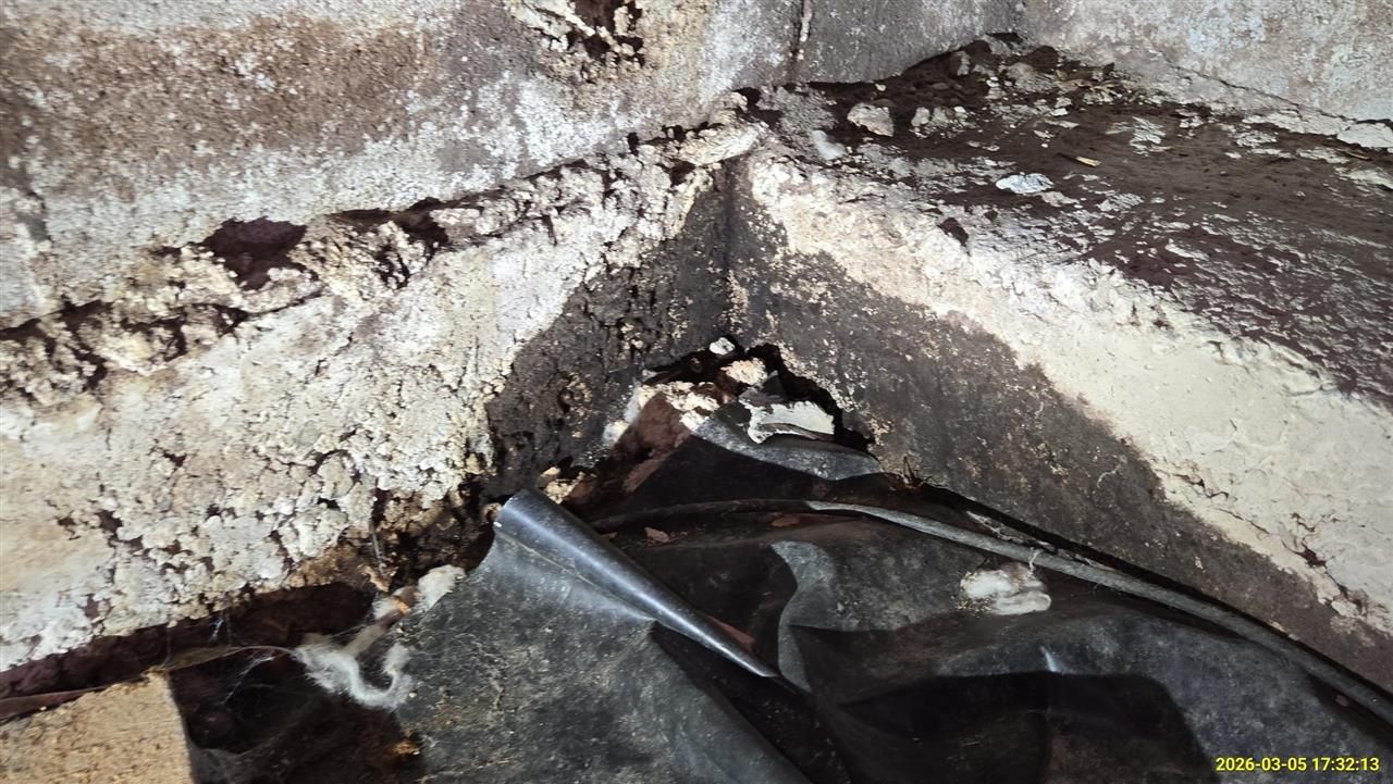 A corner where a crumbling concrete wall meets a dark, damaged floor covered by black plastic sheeting.