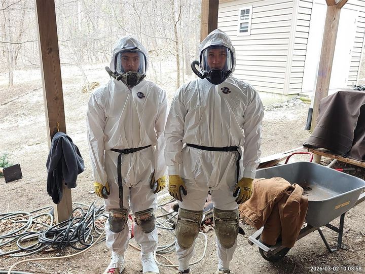 Two people wearing white protective hazmat suits and full-face respirators standing in an outdoor covered work area.