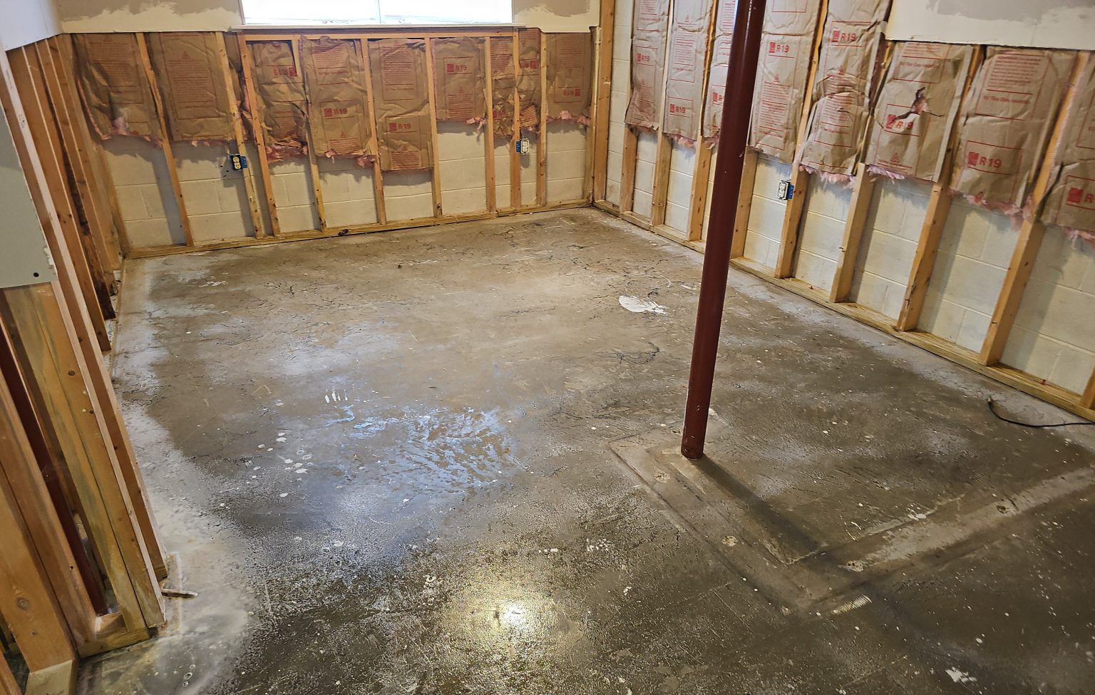 An unfinished basement featuring bare concrete floors, exposed wooden wall studs, and insulation between the studs.