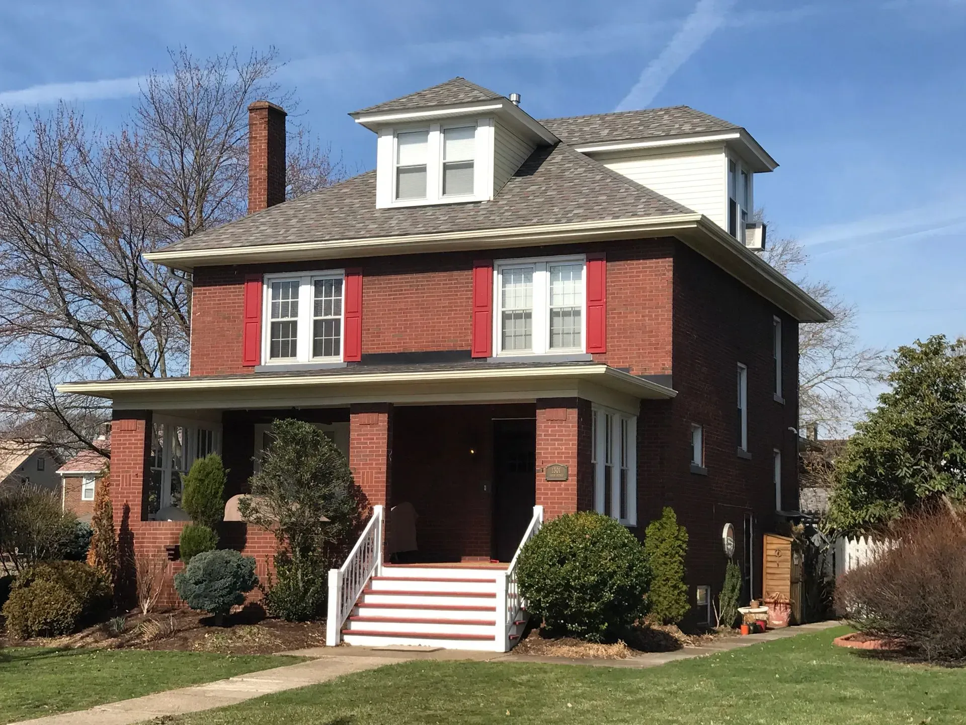 Two-story red brick house with porch, white trim, and a small yard on a sunny day.