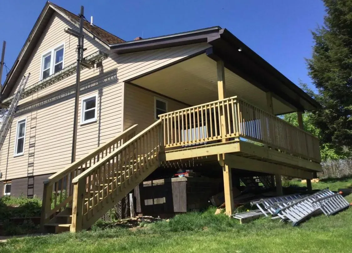 A two-story house with a wooden deck and stairs; ladders and construction materials are on the ground.