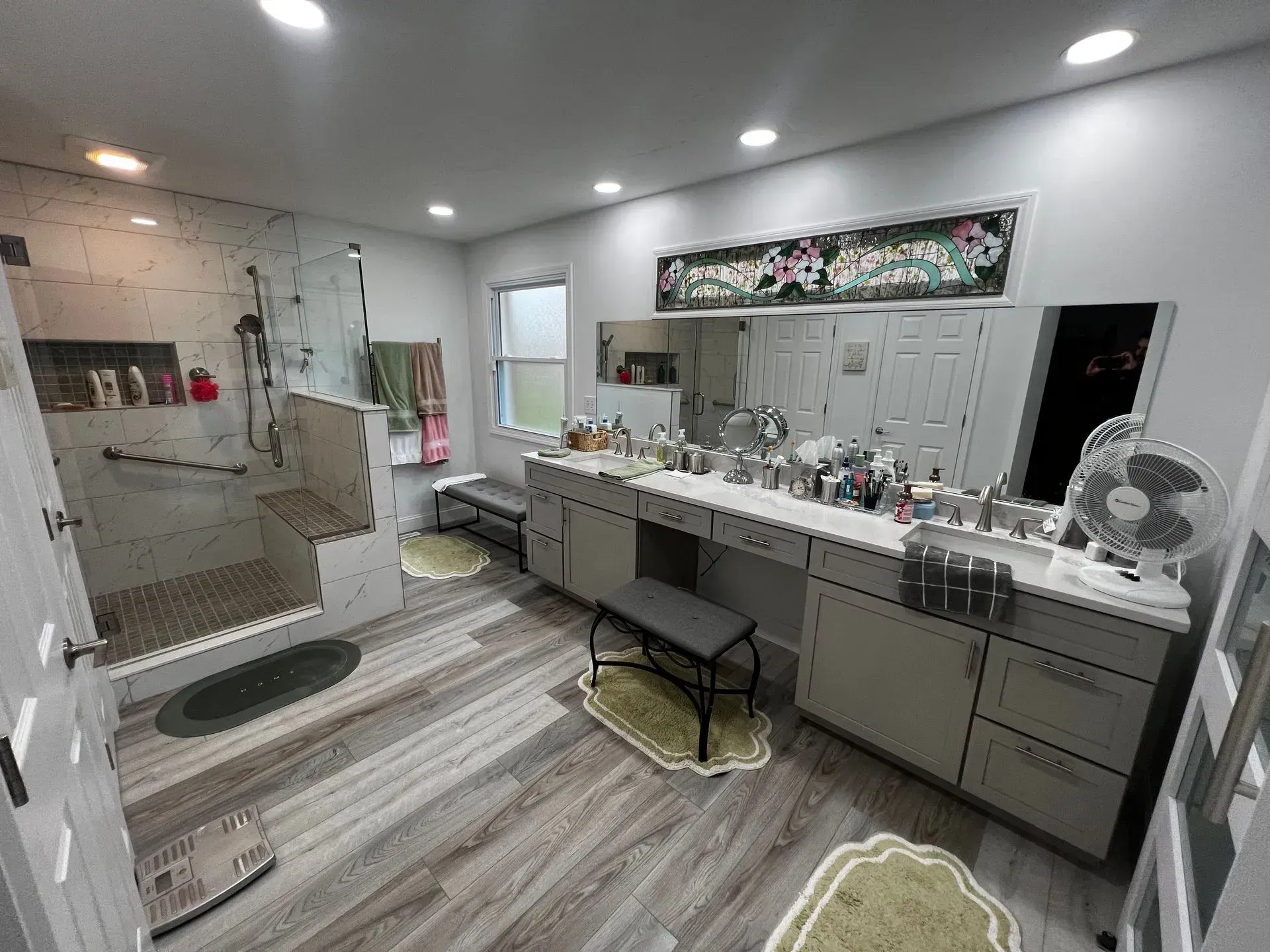 Bathroom with vanity, large mirror, shower, and gray cabinets. Light wood-look flooring and rugs.
