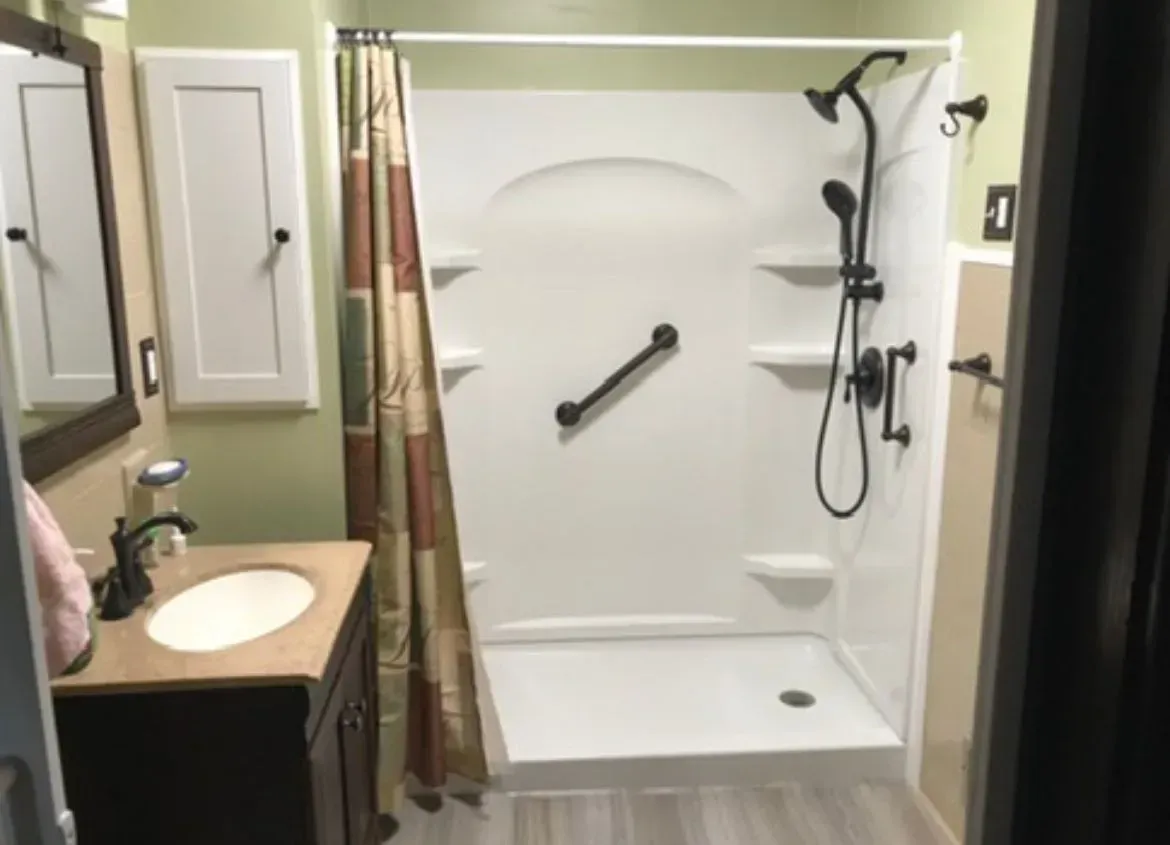 Bathroom with a white shower, grab bar, dark fixtures, and green walls. A sink is on the left.