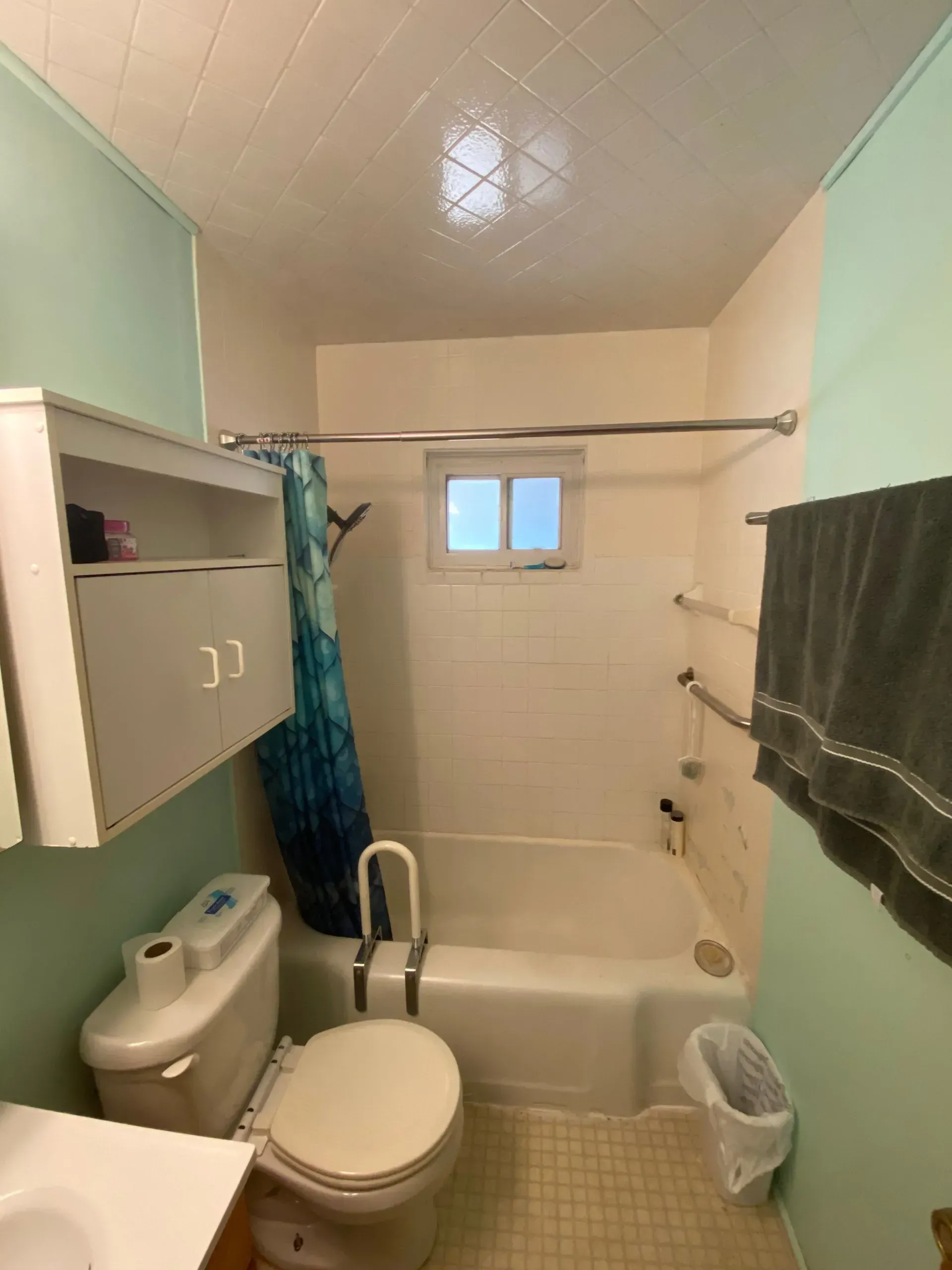 Small bathroom with tub/shower, toilet, and cabinet; light blue walls, white ceiling, and gray towel.
