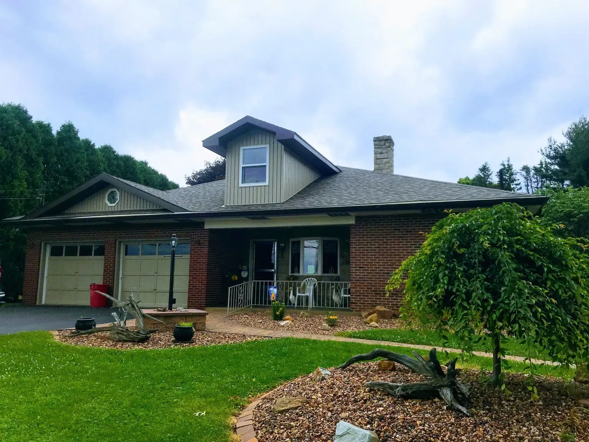 Brick house with garage, porch, and chimney, under a cloudy sky, surrounded by a lawn and small trees.