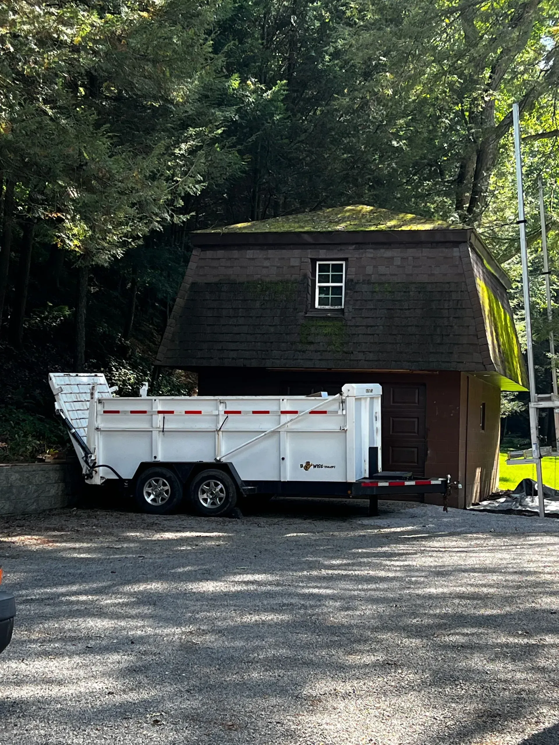 White trailer in front of a barn-style building with a mossy roof, set in a wooded area.