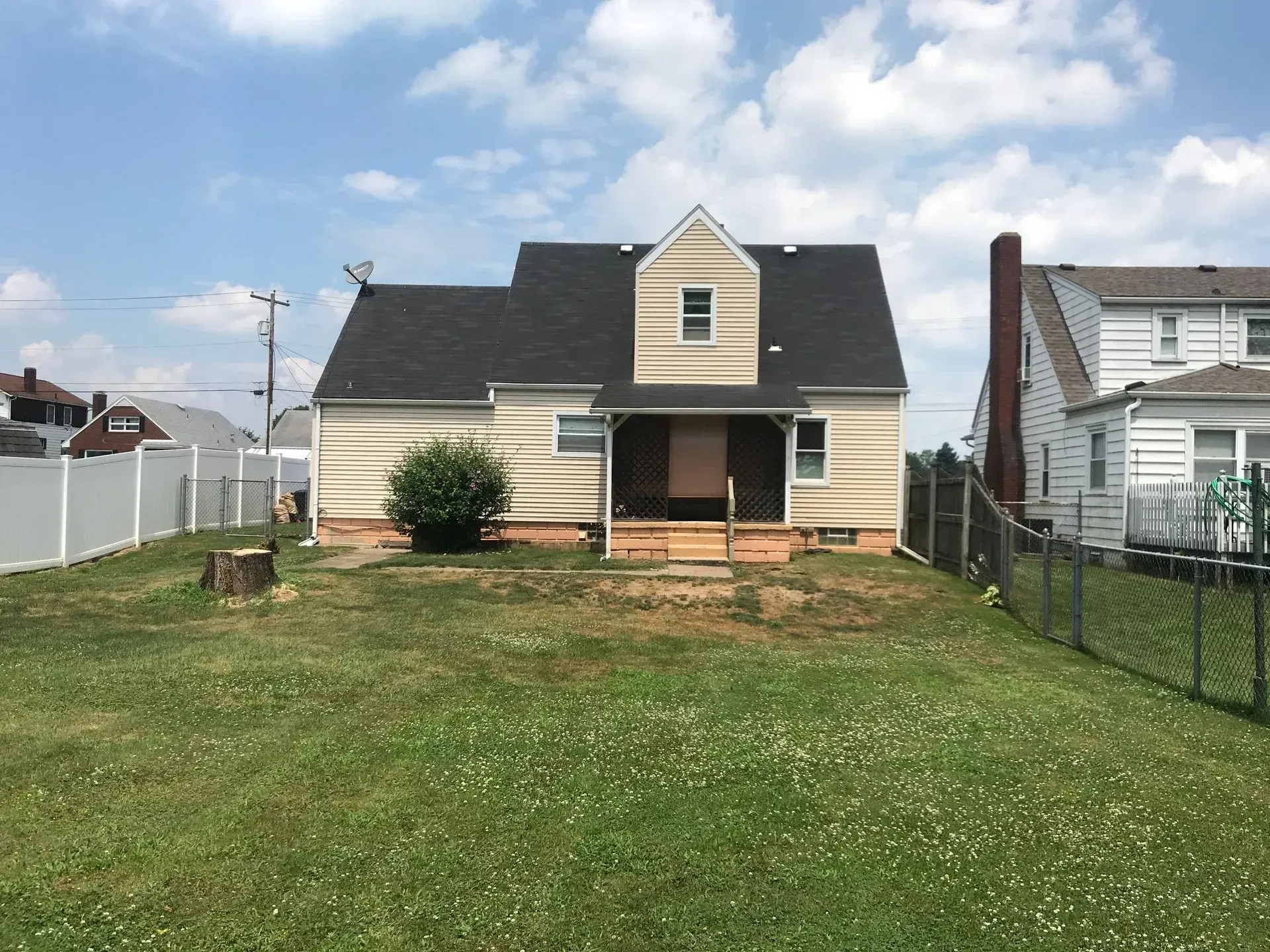 Backyard view of a beige house with a dark roof and a small porch, surrounded by a white fence and green lawn.