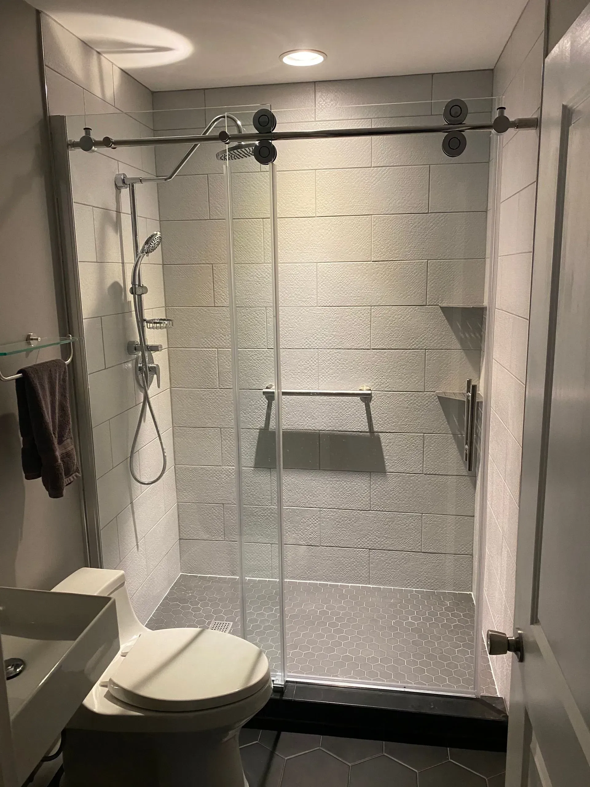 Modern bathroom with a glass shower, toilet, and sink. Silver textured tiles and a sliding glass door.