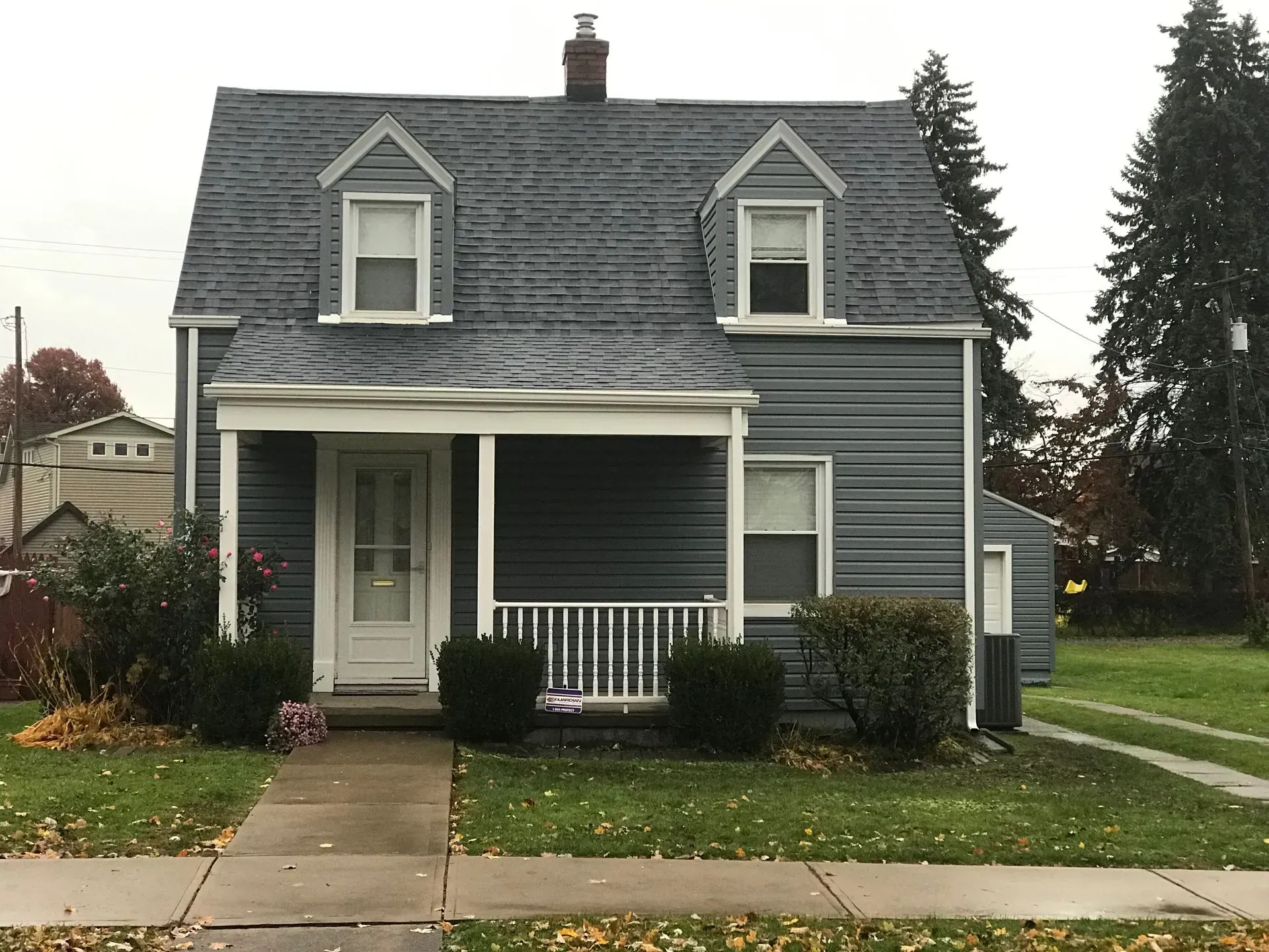 Gray two-story house with porch and dormer windows, surrounded by grass and shrubs, on a cloudy day.