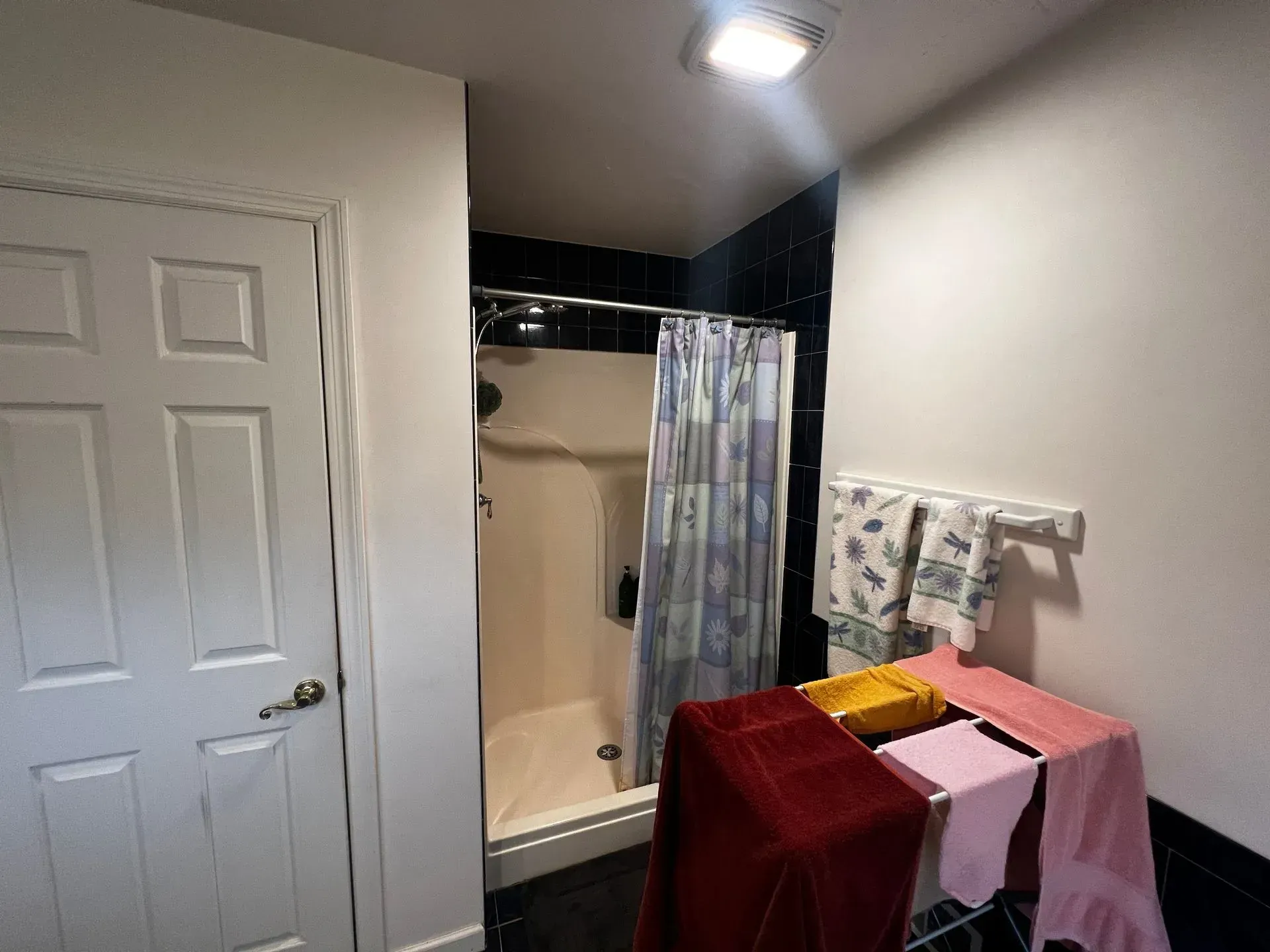 Bathroom with a white door, shower, towels hanging and folded, black tiles, and overhead light.