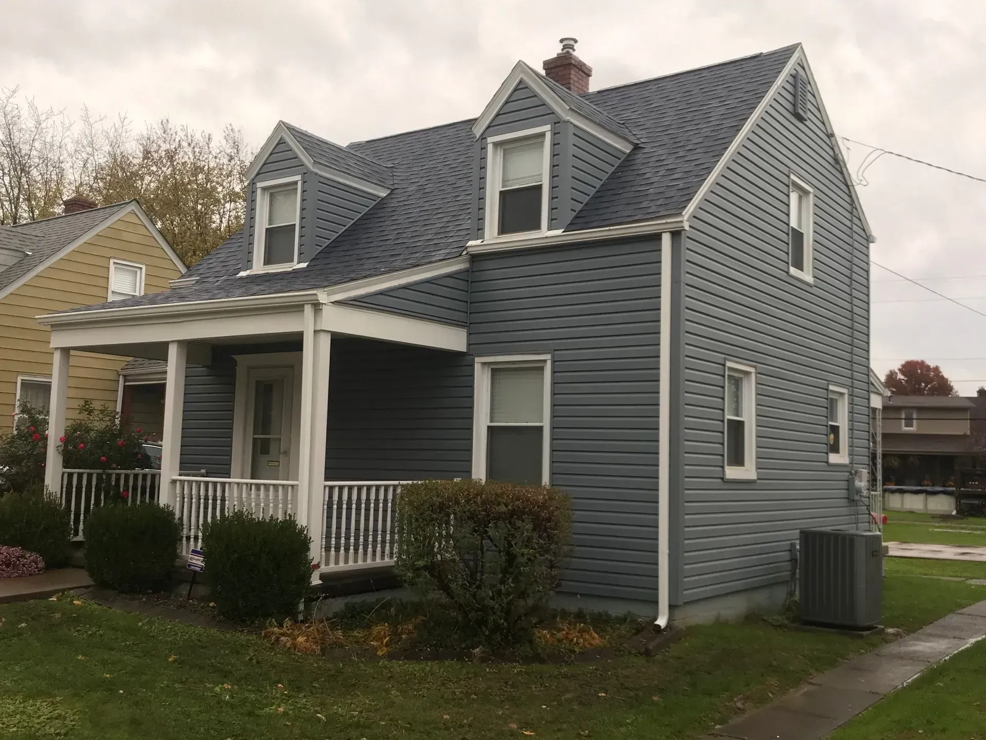 Blue-sided house with a porch and dormer windows under an overcast sky.