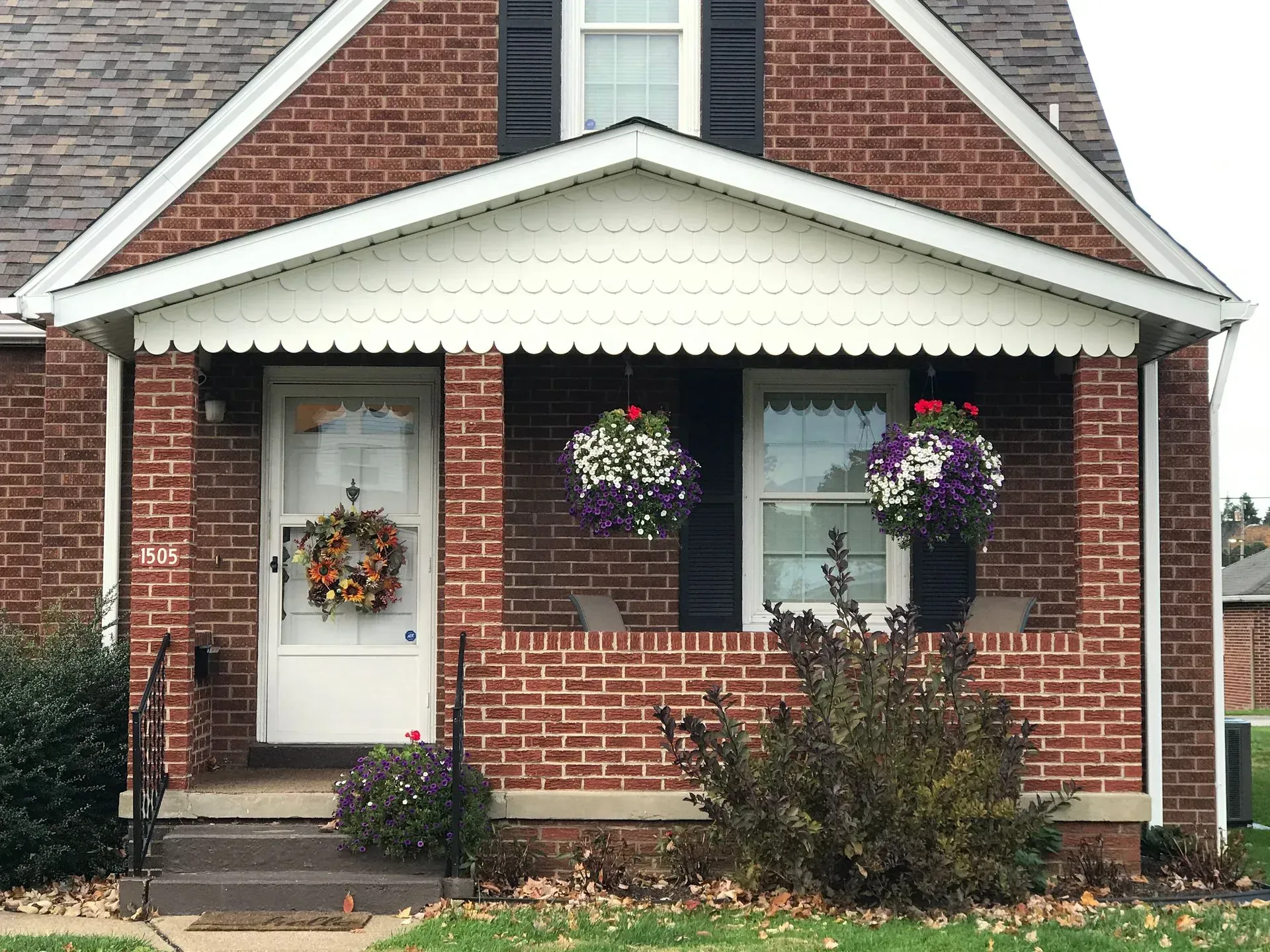 Brick house with white porch roof, door, and windows. Hanging flower baskets and fall wreath decorate the front.