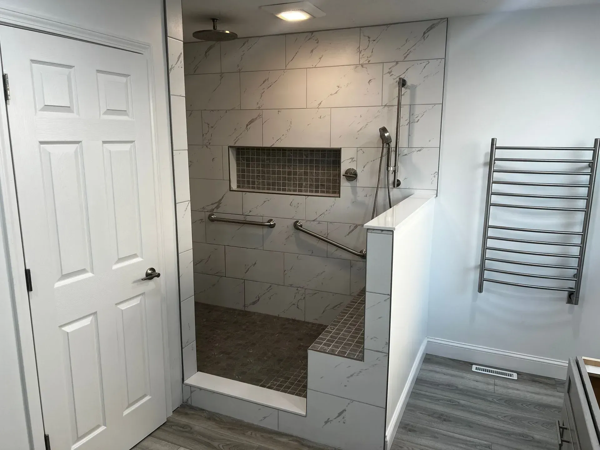 Accessible bathroom with walk-in shower. White tiled walls, shower bench, grab bars, and heated towel rack.