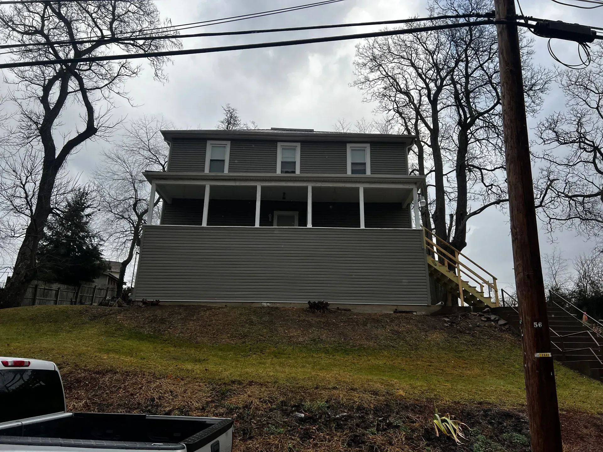 Two-story house with gray siding, porch, and a grassy hill in front. Overcast sky.