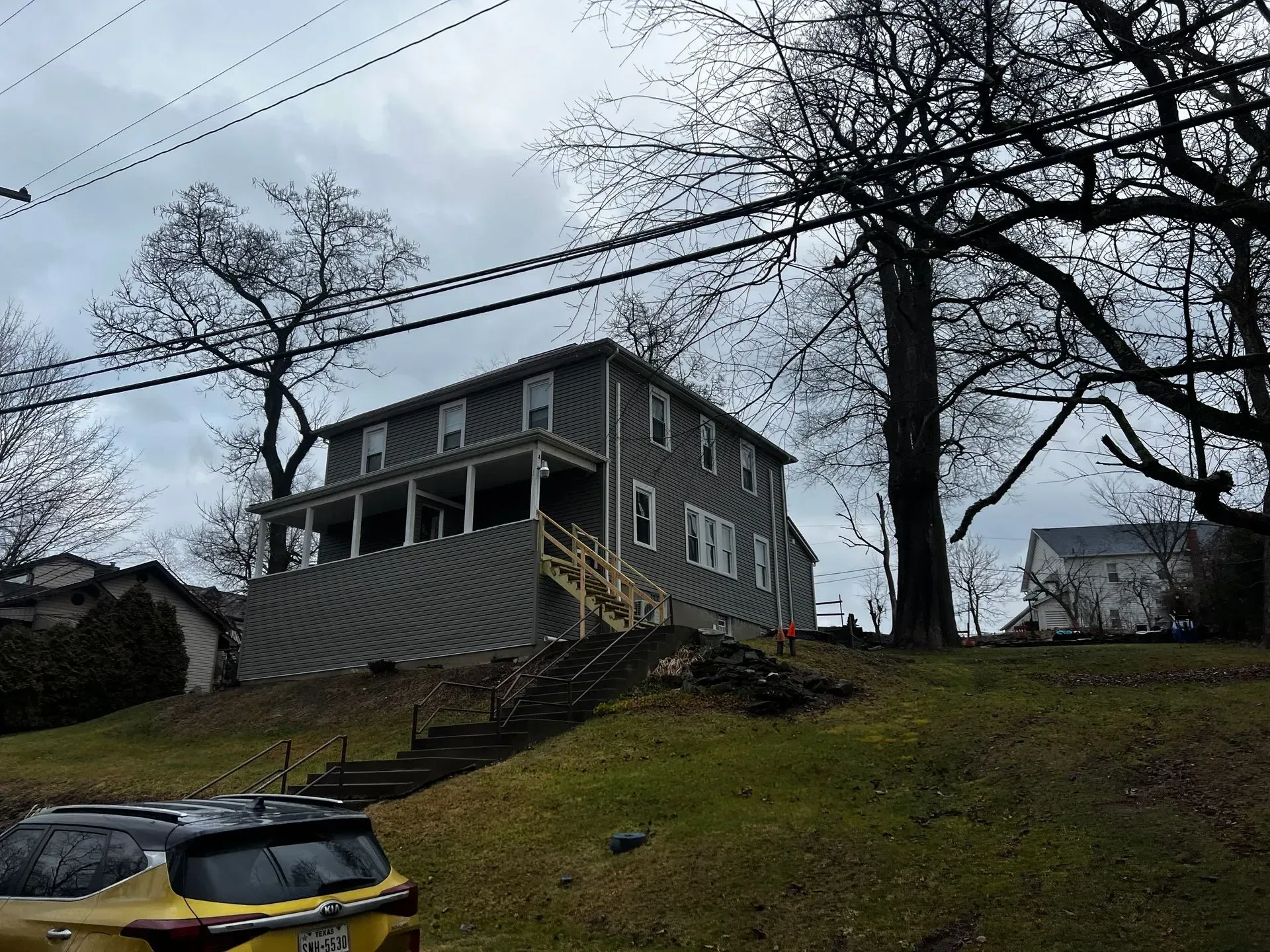 A three-story gray house on a hill, with bare trees and overcast sky. A yellow car in the foreground.