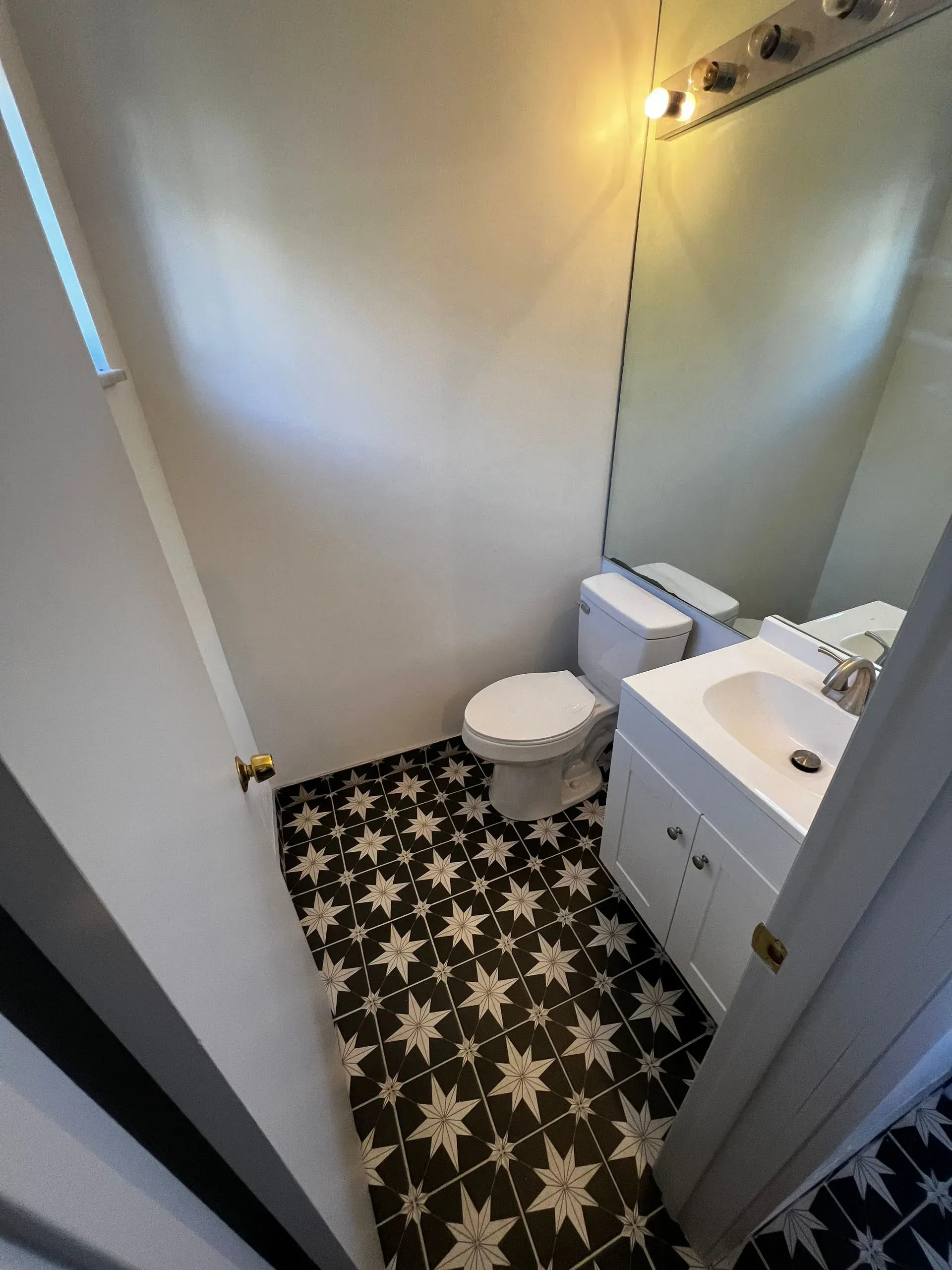 Small bathroom with star-patterned black and white floor tiles, toilet, white sink, and large mirror.