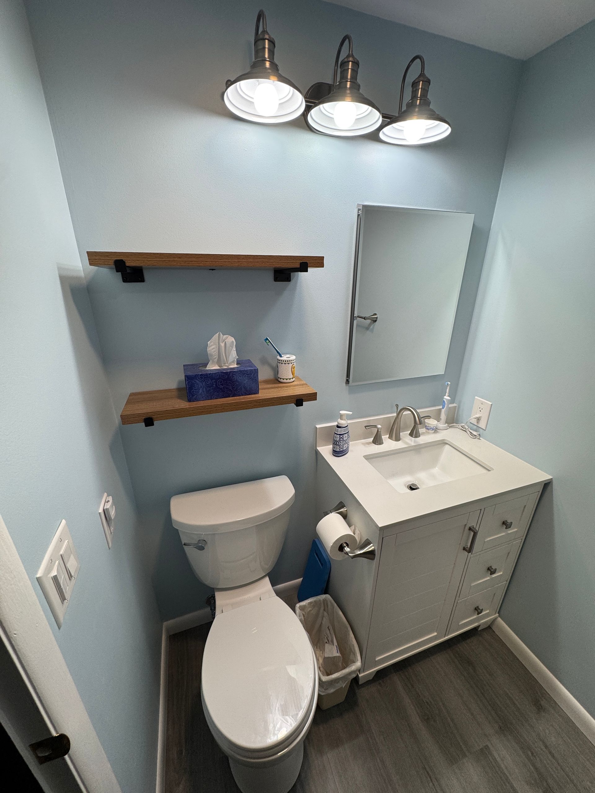 Small bathroom with toilet, sink, mirror, shelves, and overhead lights. Light blue walls, gray floors.