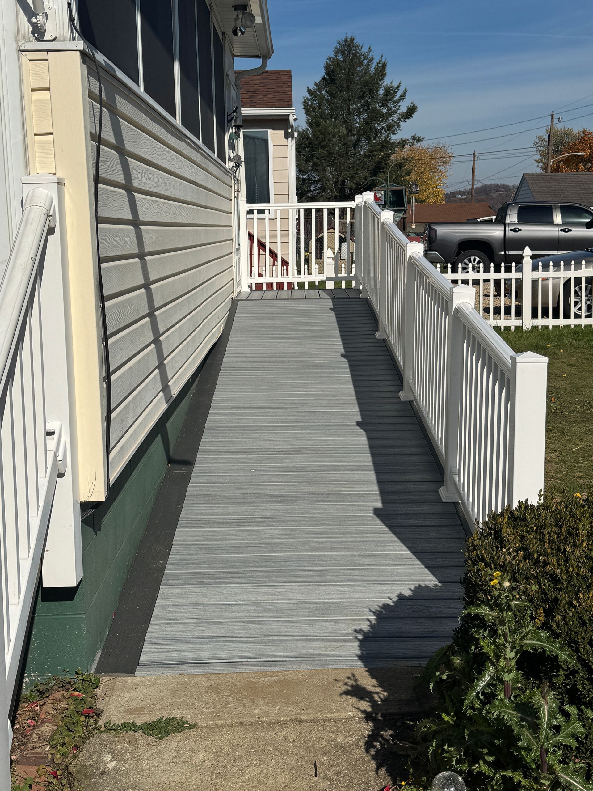 Gray ramp with white railings leads to a house entrance. Green grass and a white fence are in the background.