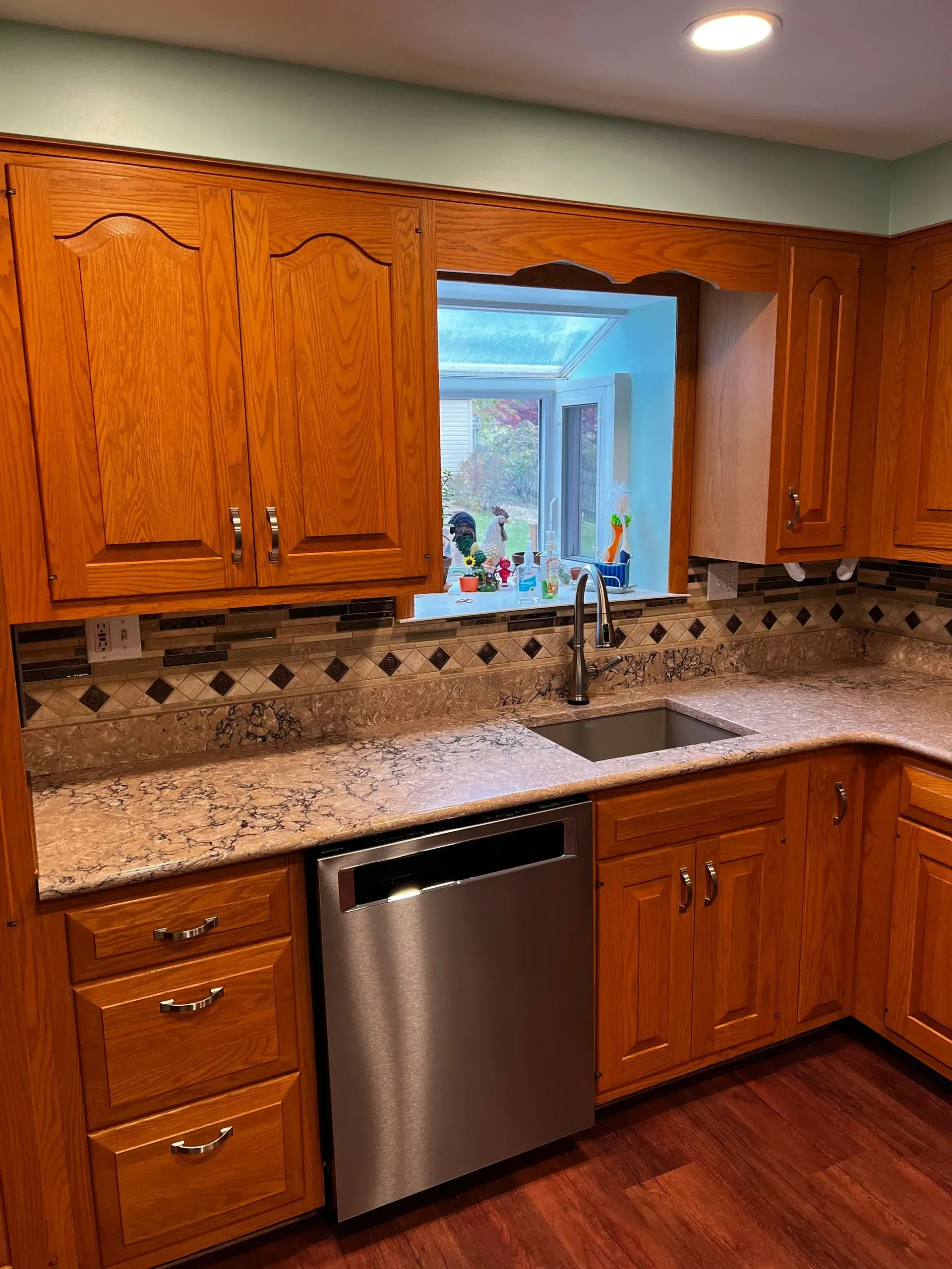 Kitchen with wooden cabinets, granite countertop, and stainless steel dishwasher. Decorative backsplash, view through window.