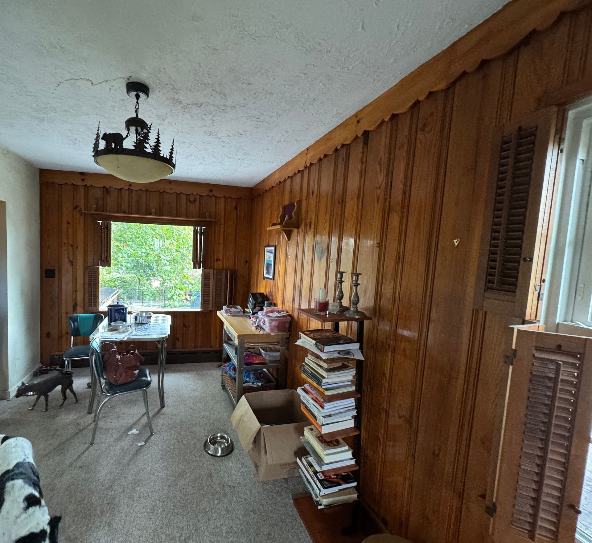 A room with wood-paneled walls, a table, window, and a dog.  Books and various items are on the table and floor.