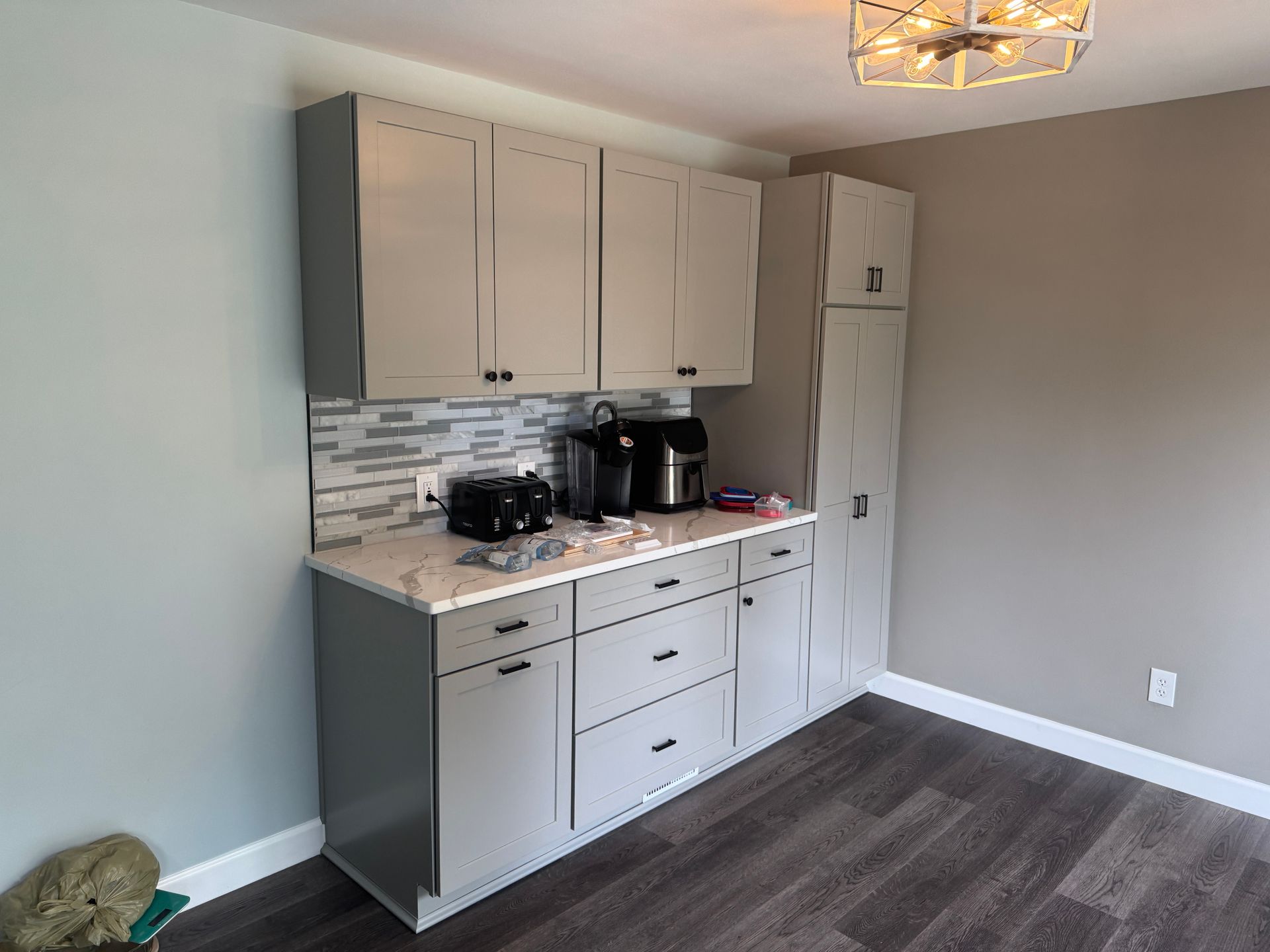 Gray cabinets in a room with a coffee station. White countertops and backsplash, dark hardware.