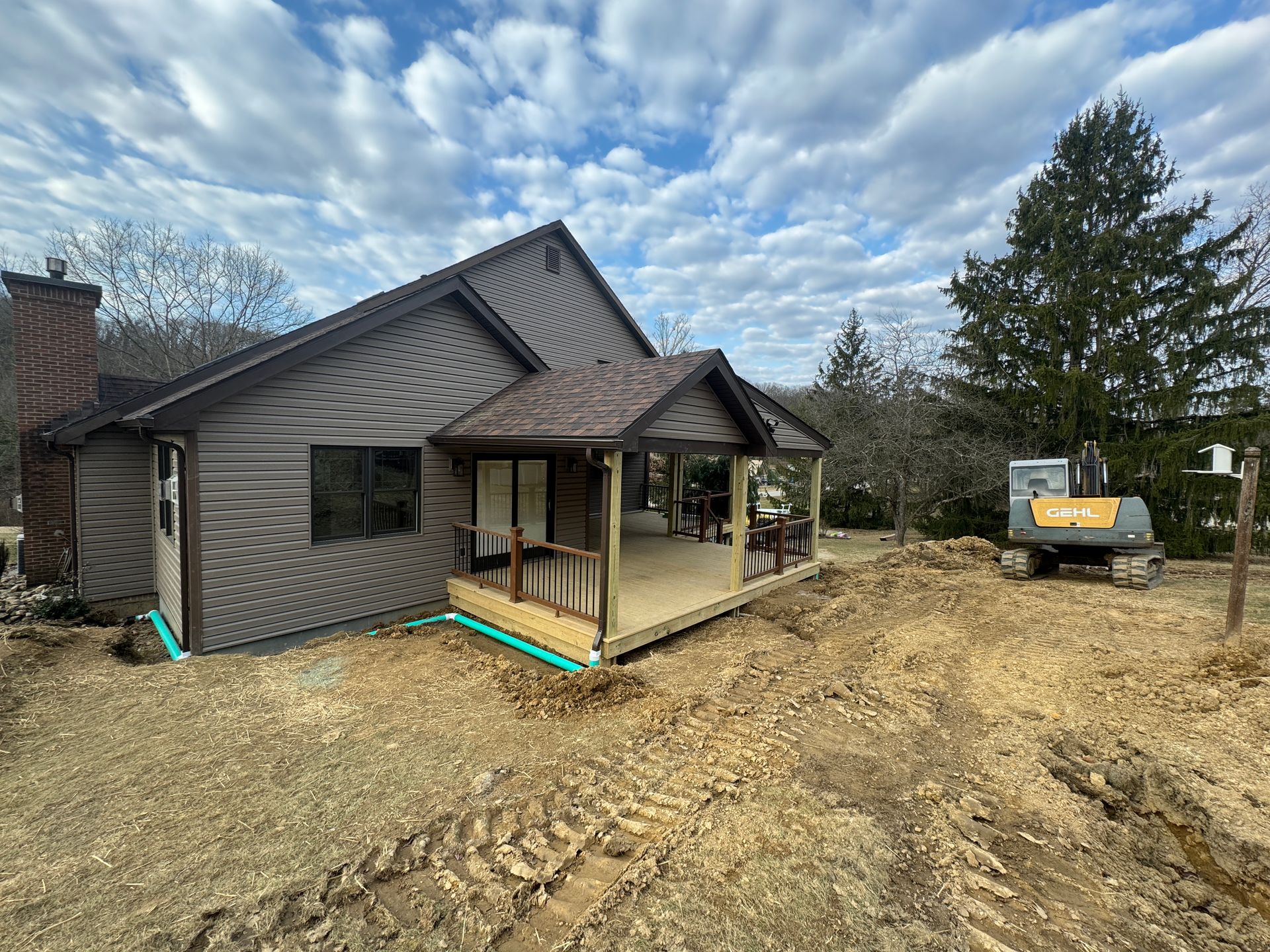 House with brown siding, porch under construction, excavator on dirt. Cloudy sky.