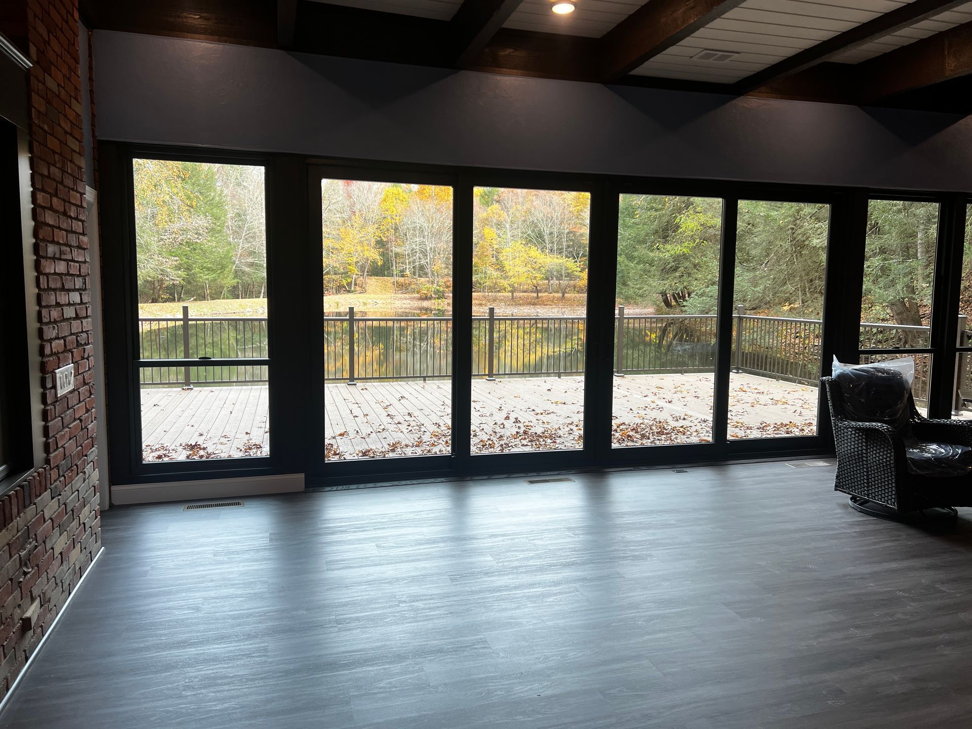 Room with large windows overlooking a lake, brick wall, dark wood beams, and a black chair.