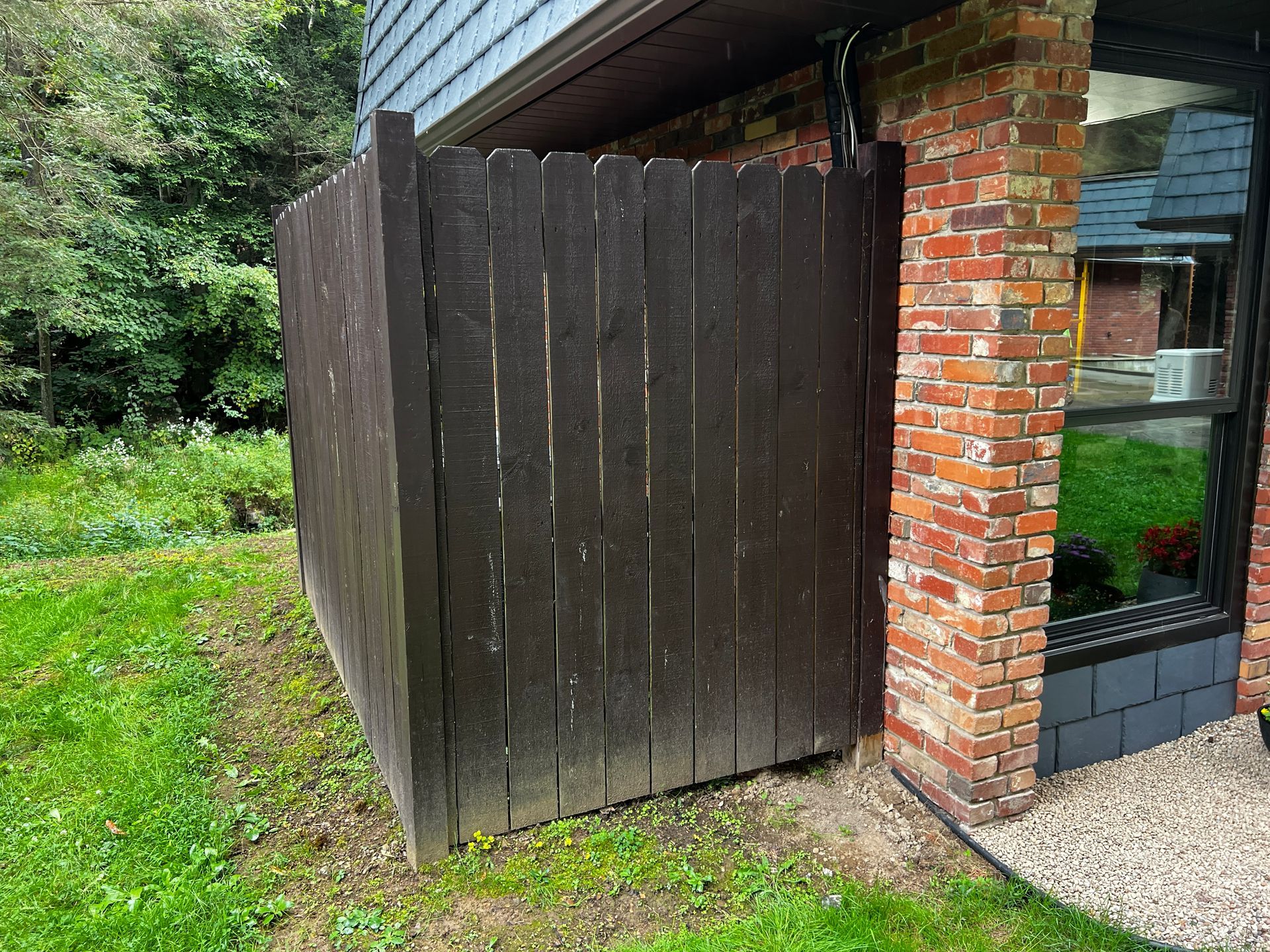 Dark brown wooden fence next to a brick building with a window, on green grass.