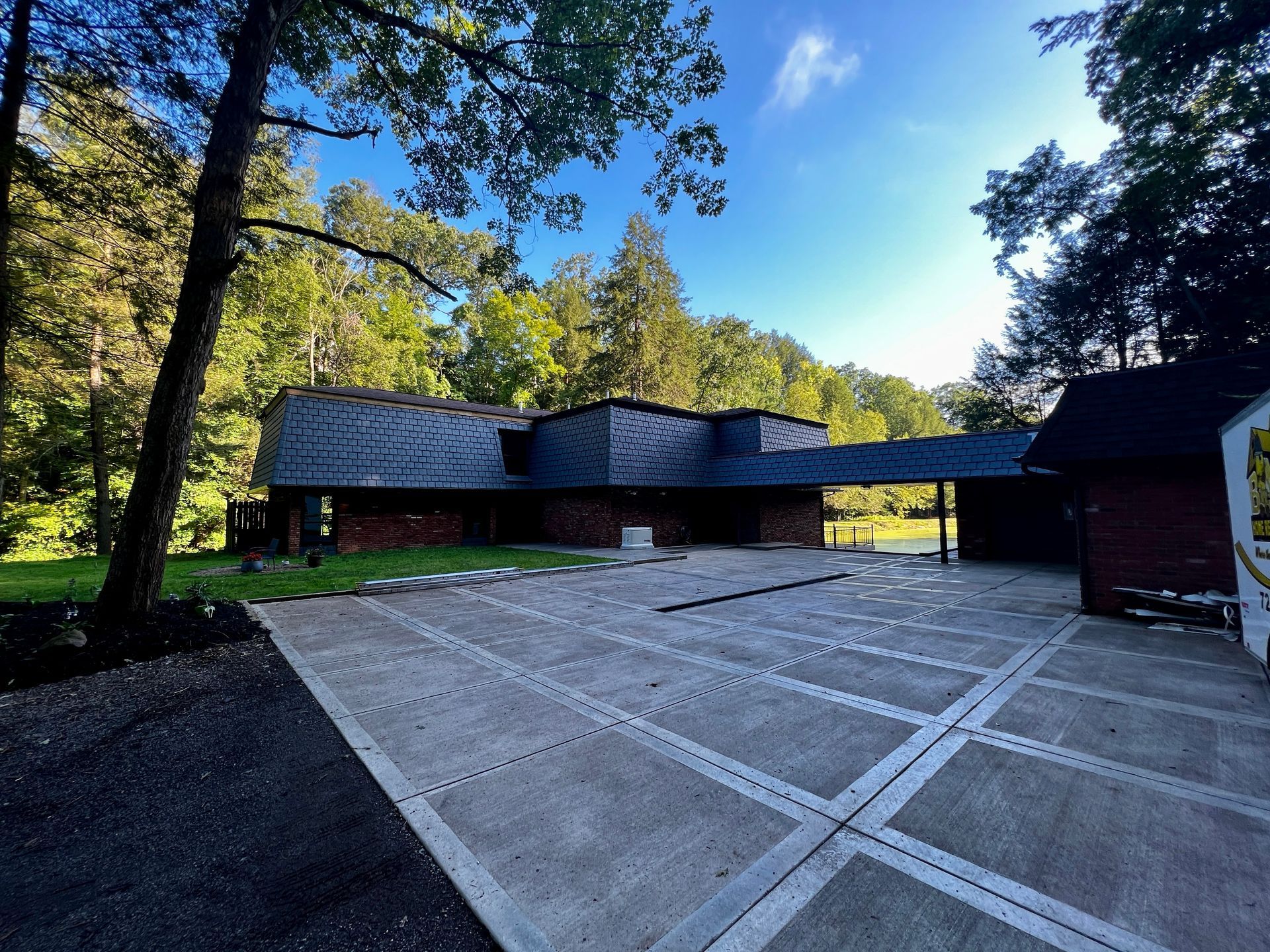 House with dark roof, brick walls, and concrete driveway surrounded by trees on a sunny day.