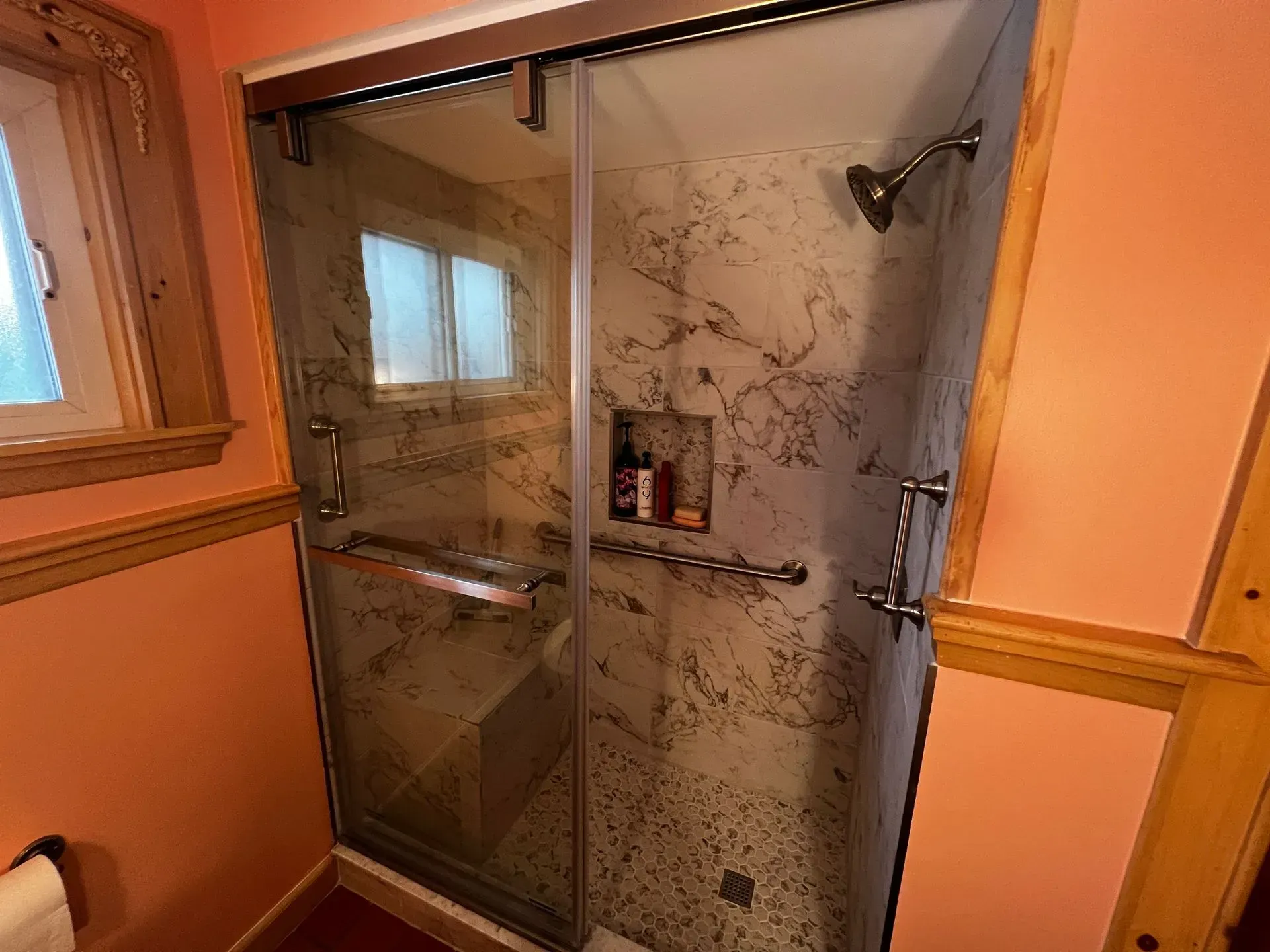 Shower with glass doors and marble-look tile. Includes shower head, built-in shelving and grab bars.