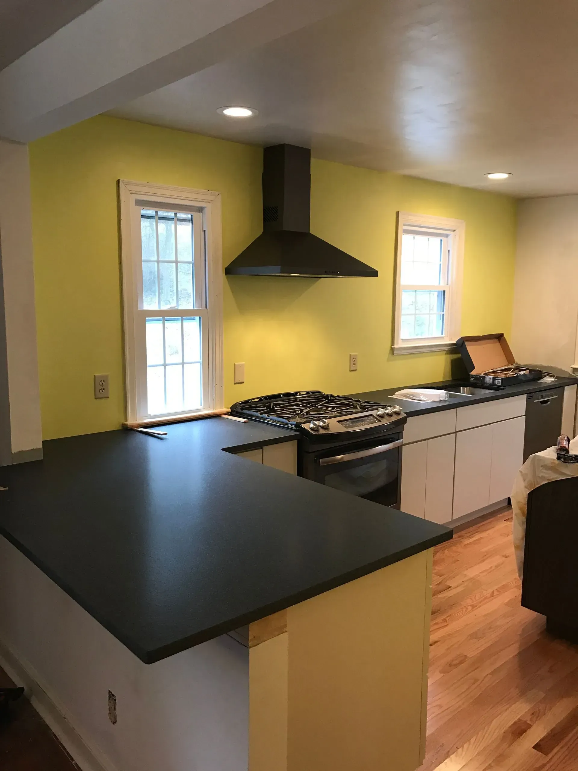 Kitchen with yellow walls, black range hood, and dark countertops. White cabinets and wood floors.