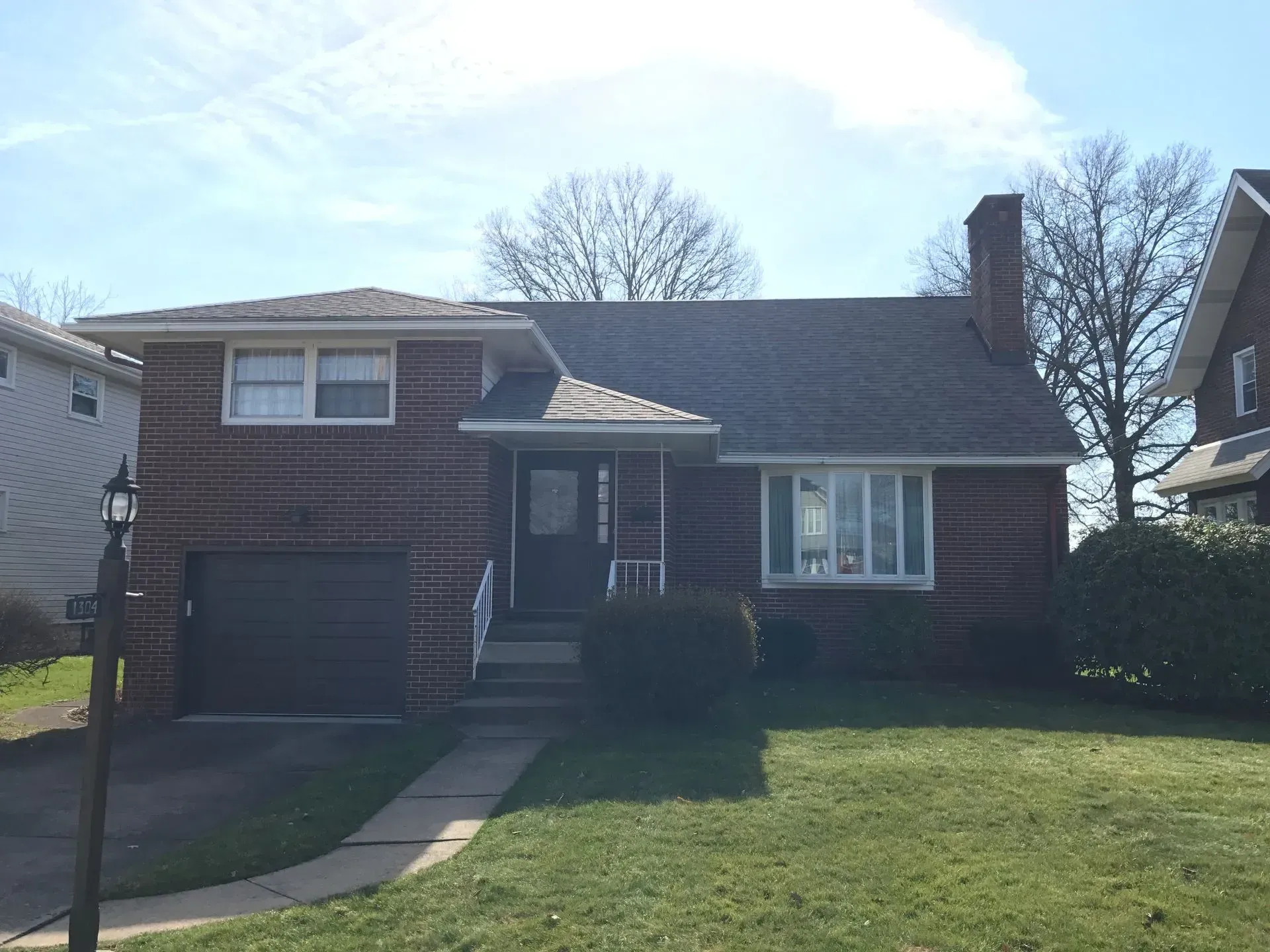 Brick house with attached garage, chimney, and front porch. Green lawn with tree and overcast sky.