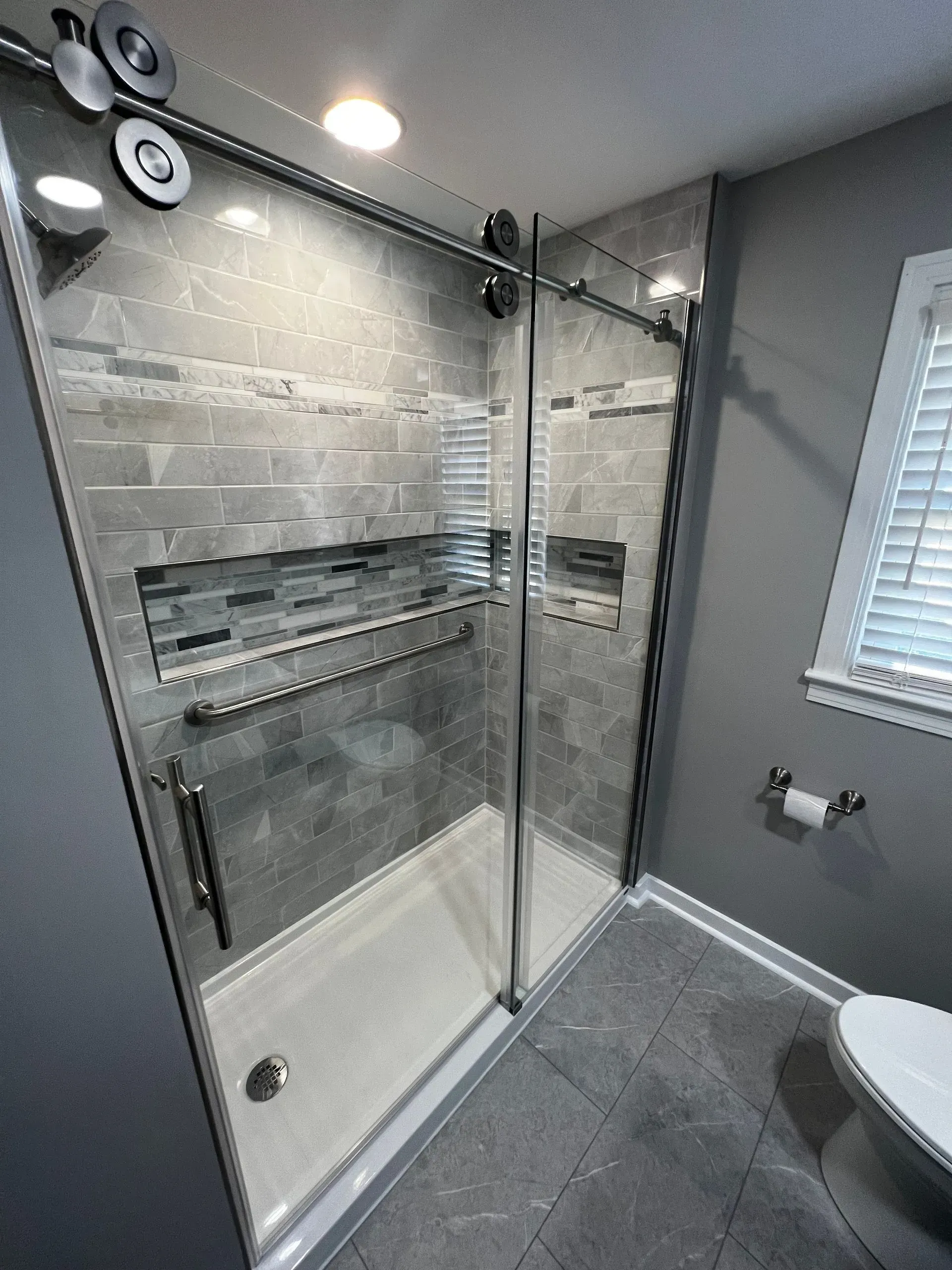 Modern shower with gray tiled walls, glass doors, and a built-in shelf.  A toilet is to the right.