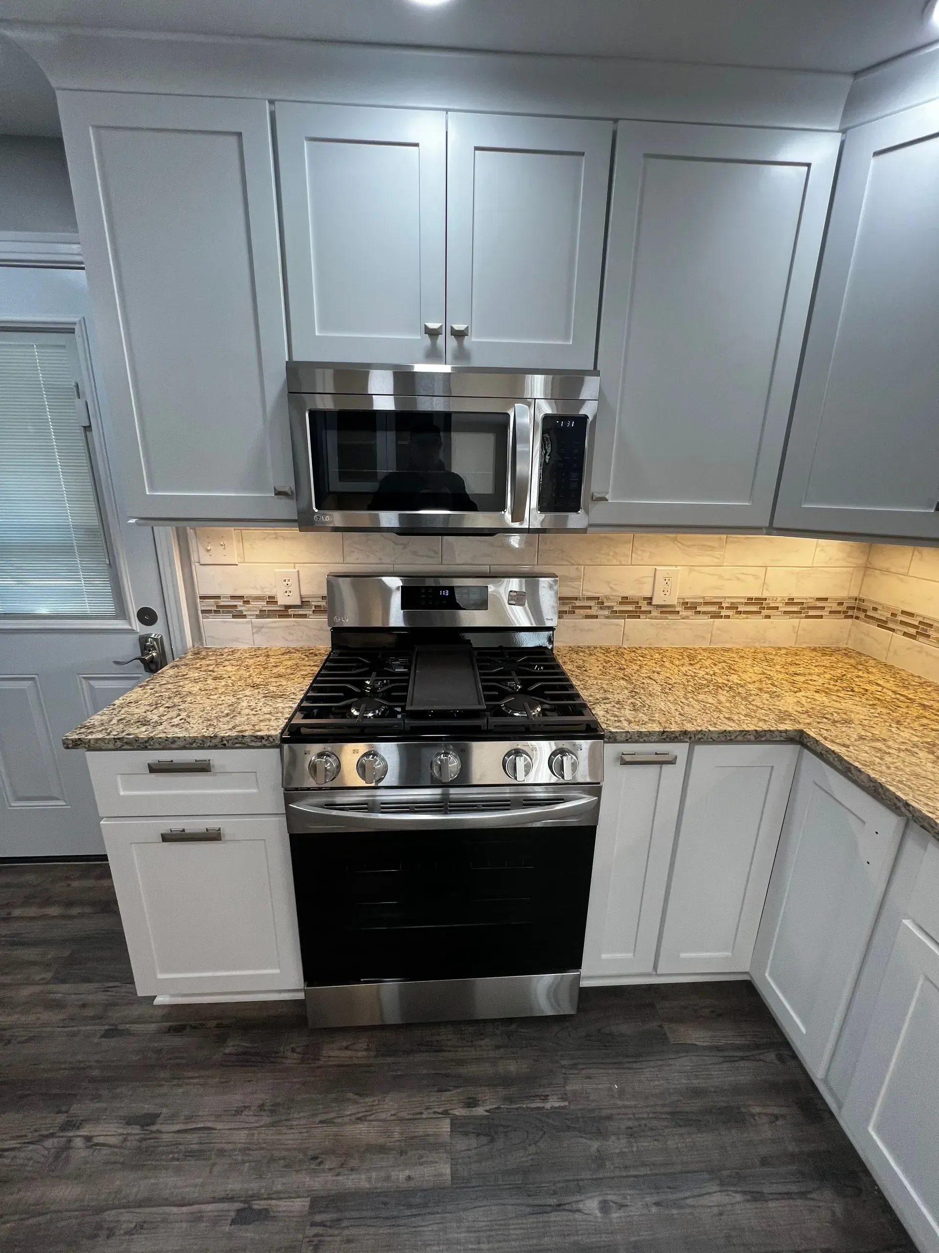 A modern kitchen with grey cabinets, granite countertops, stainless steel appliances, and dark wood-look flooring.