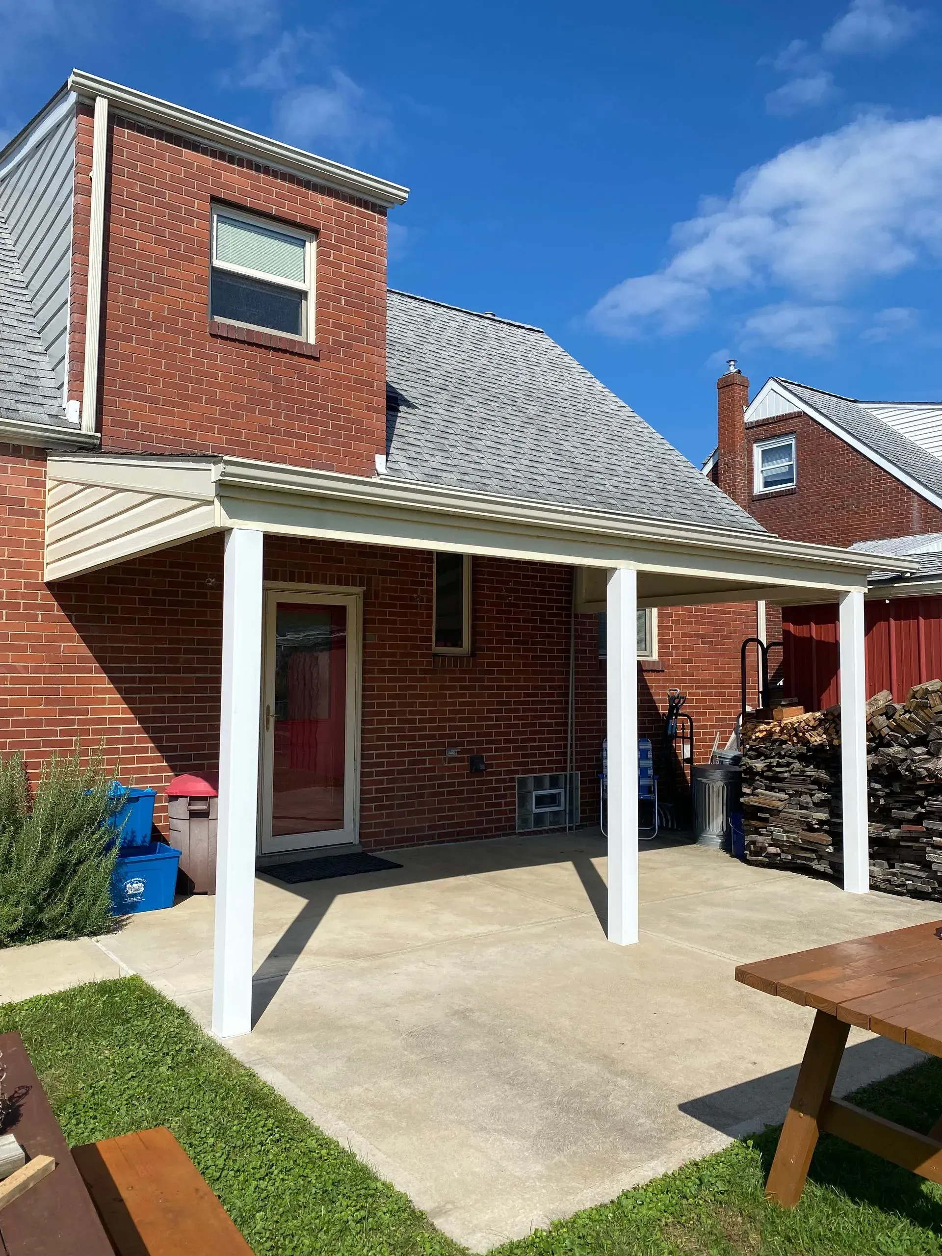 Backyard patio with red brick house, white pergola, concrete patio, firewood, blue sky.