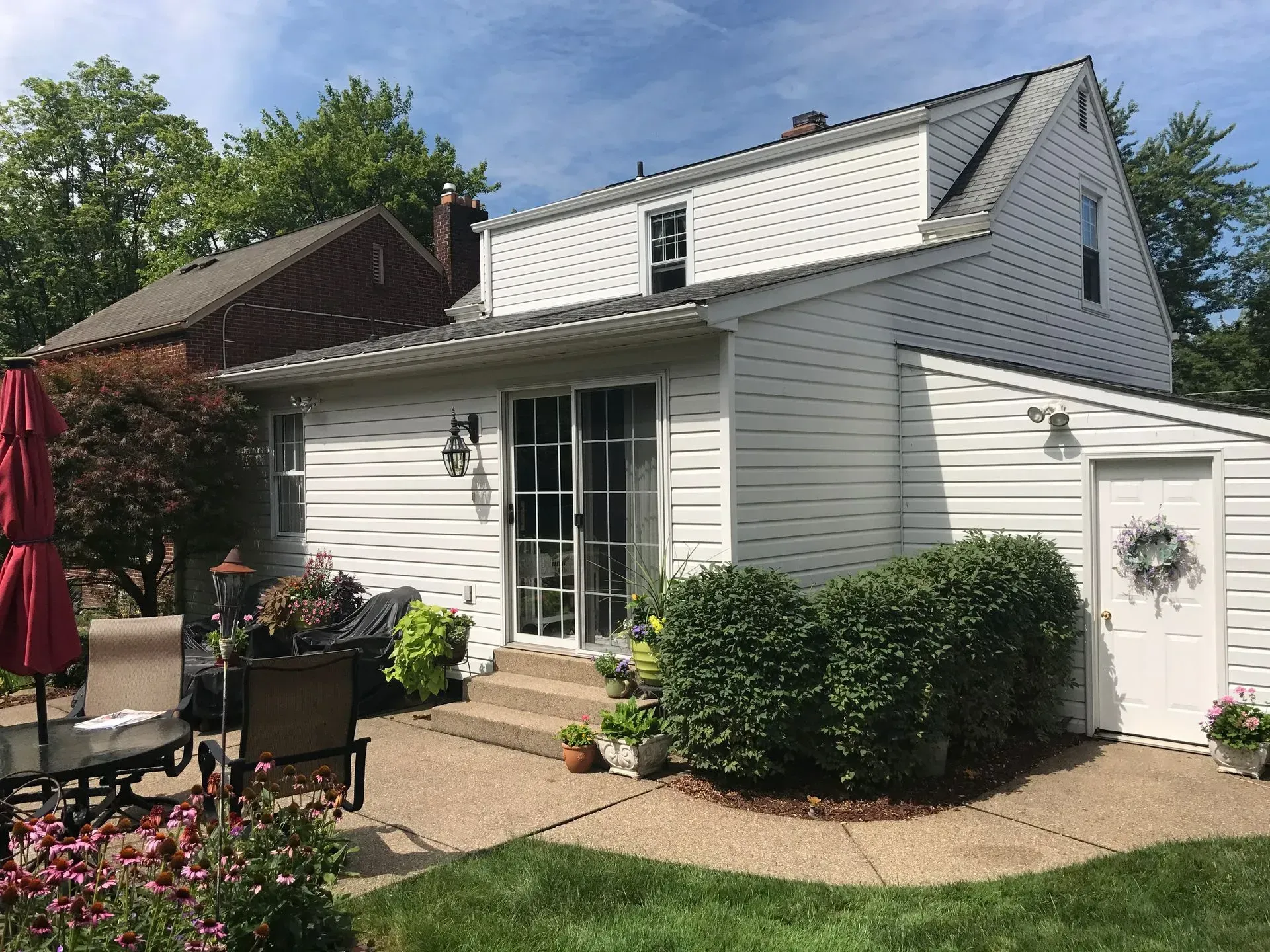 White two-story house with a small shed, a sliding glass door, and a concrete patio; surrounded by greenery and plants.